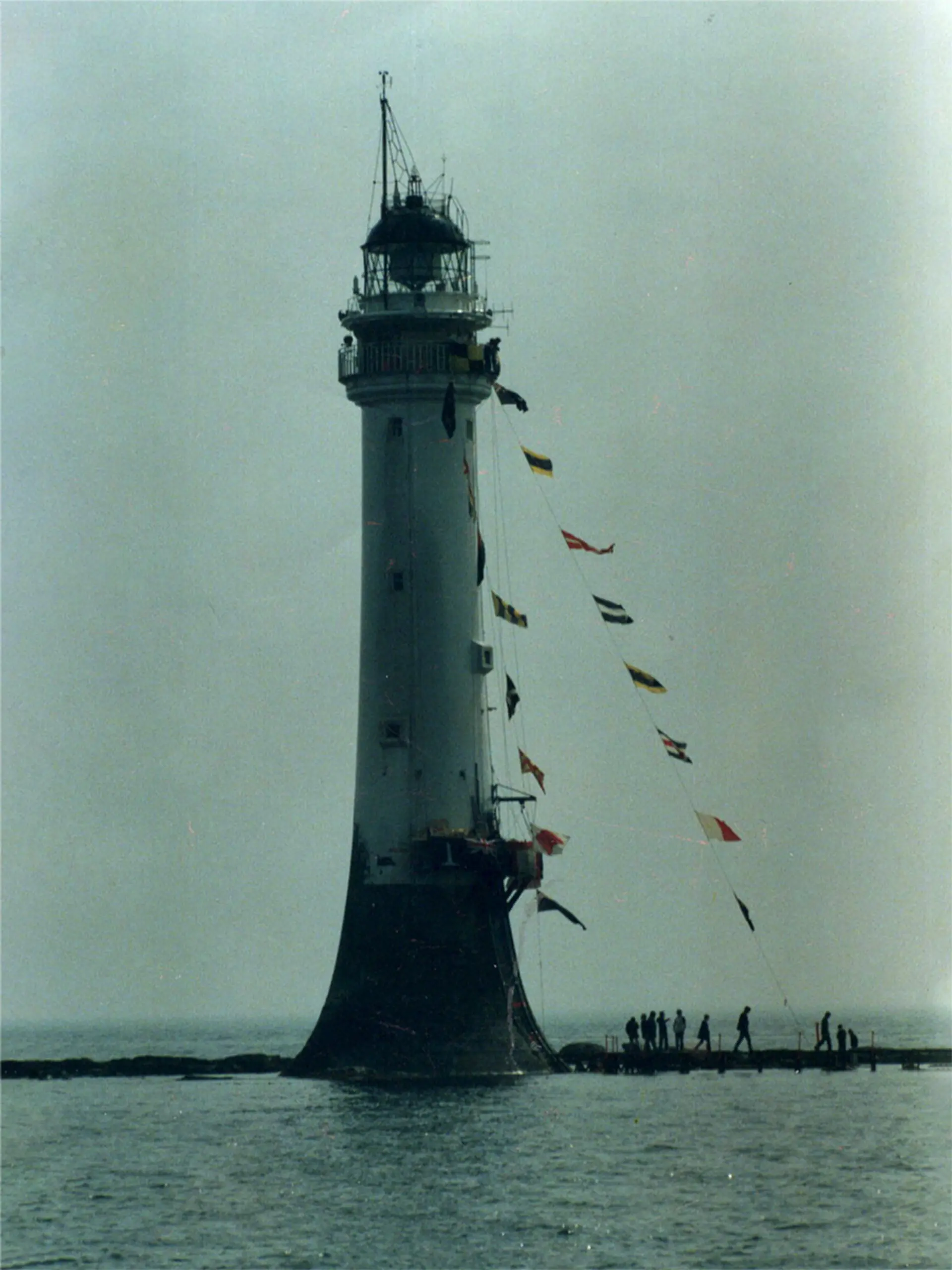 An image of The Bellrock Lighthouse in Arbroath. The lighthouse is centred in the picture and has flags attached from the top to the ground. There are people in the bottom right hand corner of the image, they look tiny. 