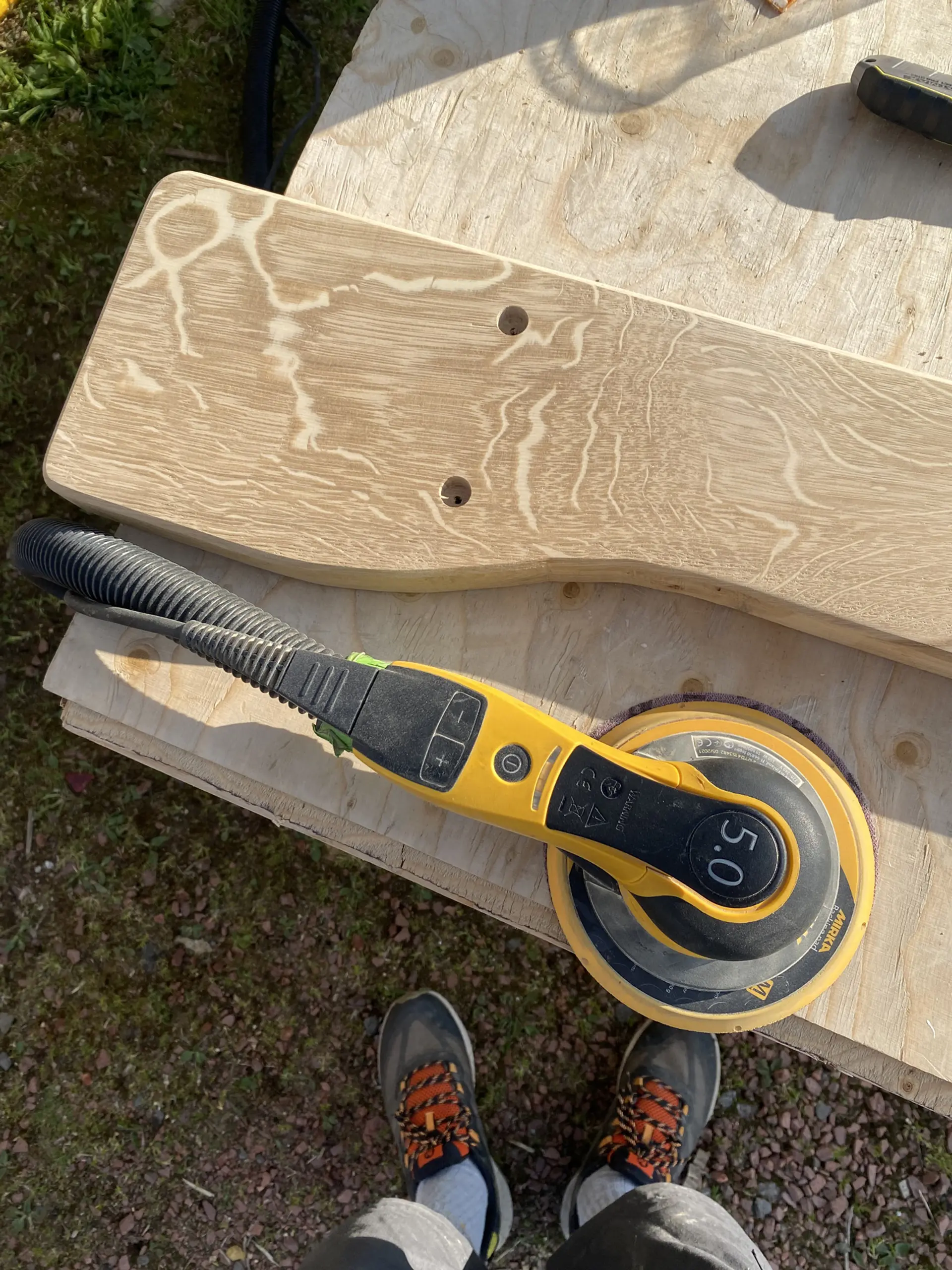 A sanded plank of oak, with 2 holes drilled in one section, sits outside. A pair of feet in the shot. The plank is pictured close up and we can see patterns, like water ripples, in the grains of the wood. What looks like a small, electric sander, sits on it. 