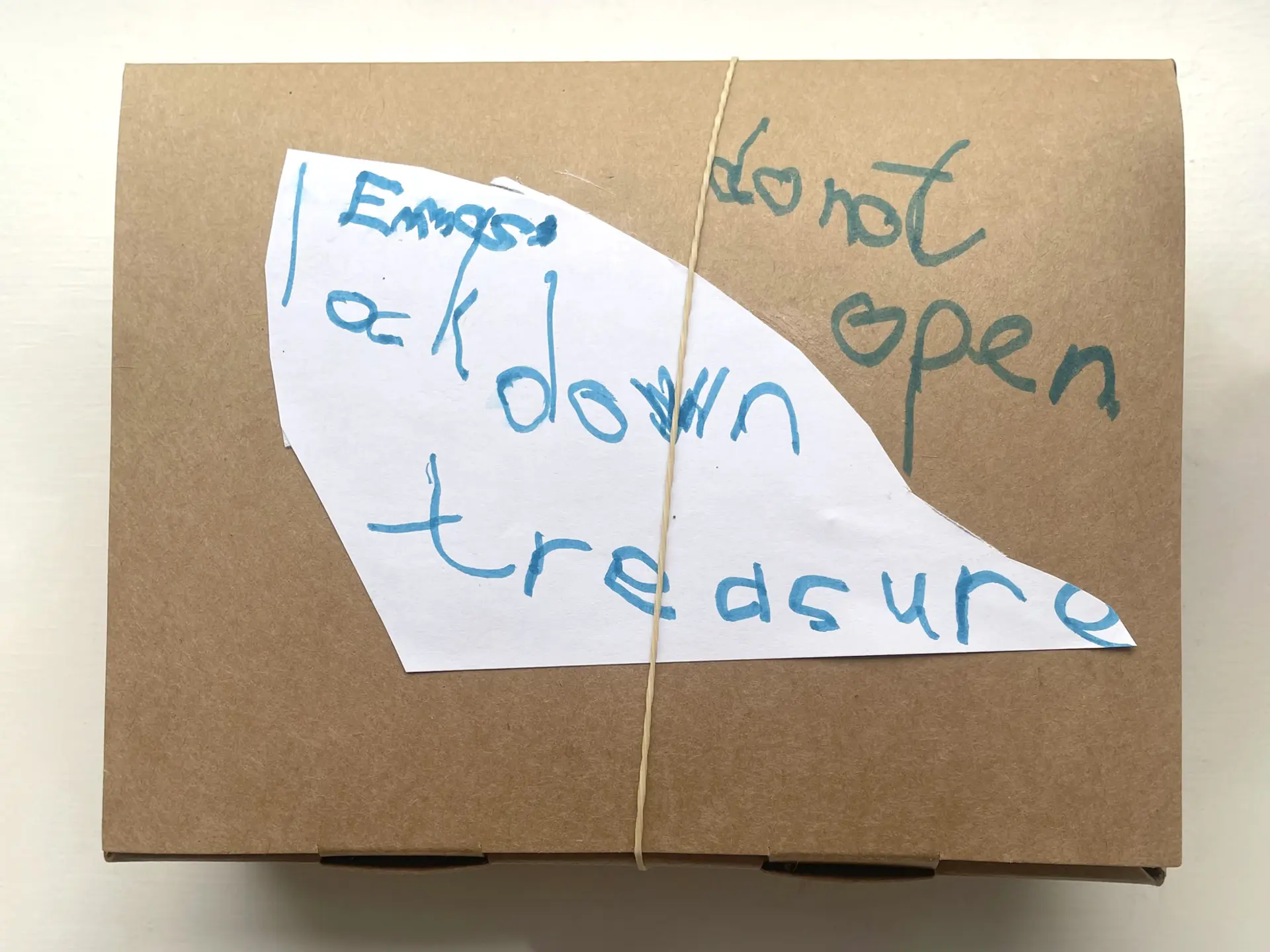 Emma’s lockdown treasures  A brown cardboard box with an elastic band around it. Written in blue felt tip pen it says ‘Emmas’s lockdown treasure, do not open’