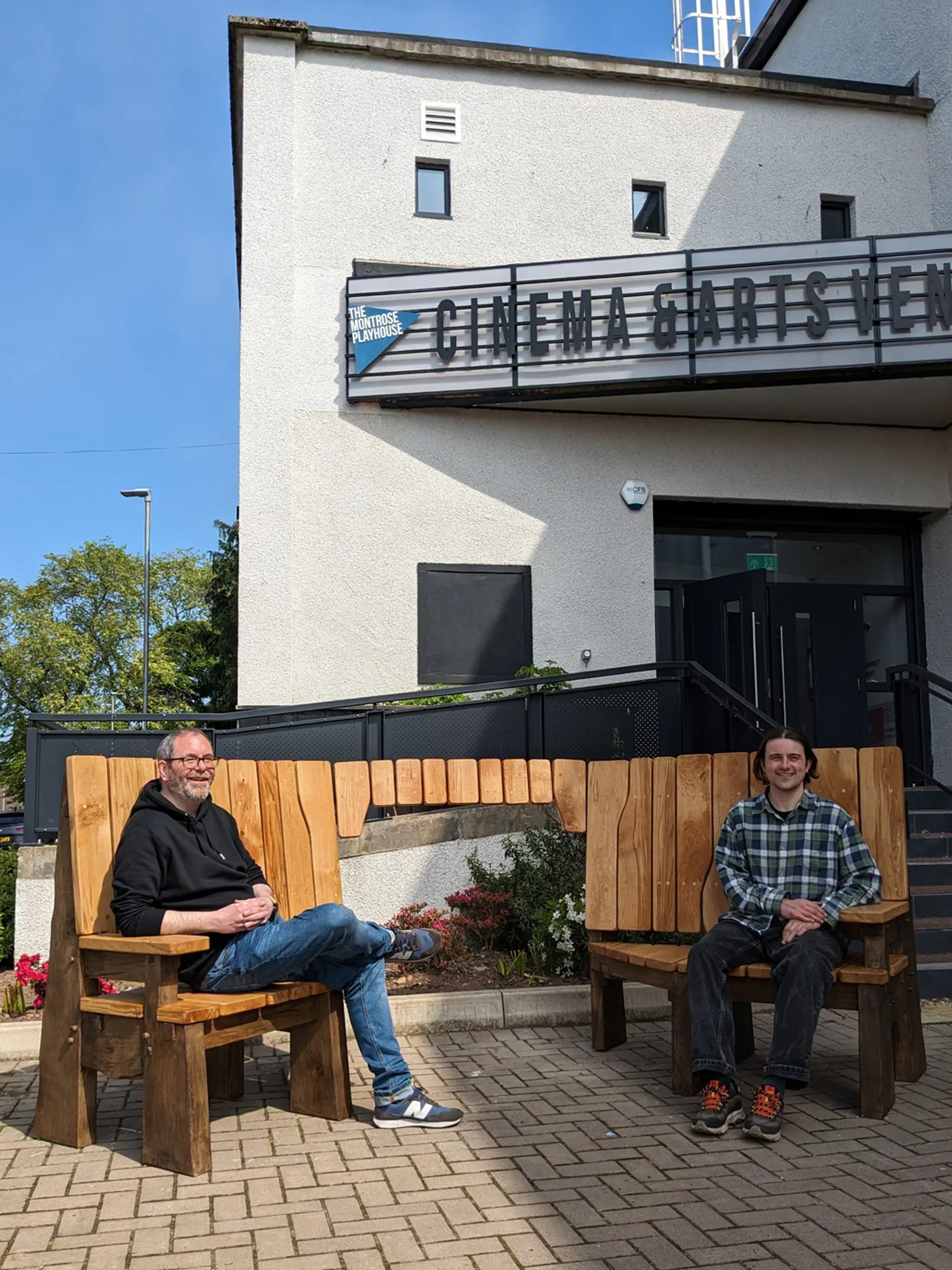 A sunny blue sky behind a white building that says, ‘Cinema and Arts Venue’. There is a solid wooden bench, two men sit at opposite ends of the bench, both are smiling at the camera.