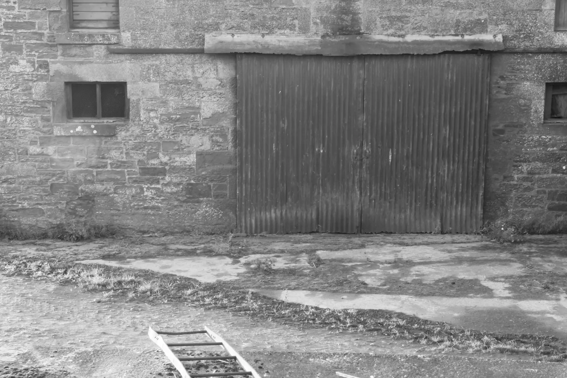 A grayscale image of a farm building. The ground is made of large stone slabs, and we are facing a stone facade of a building. The door is corrugated and there are two small windows. 