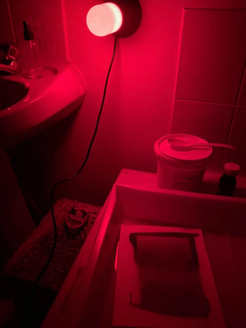 A moody red photograph showing a small section of a darkroom; a light attached to the wall beside a ceramic sink. An analogue print is being processed, floating in a small tank of solution.