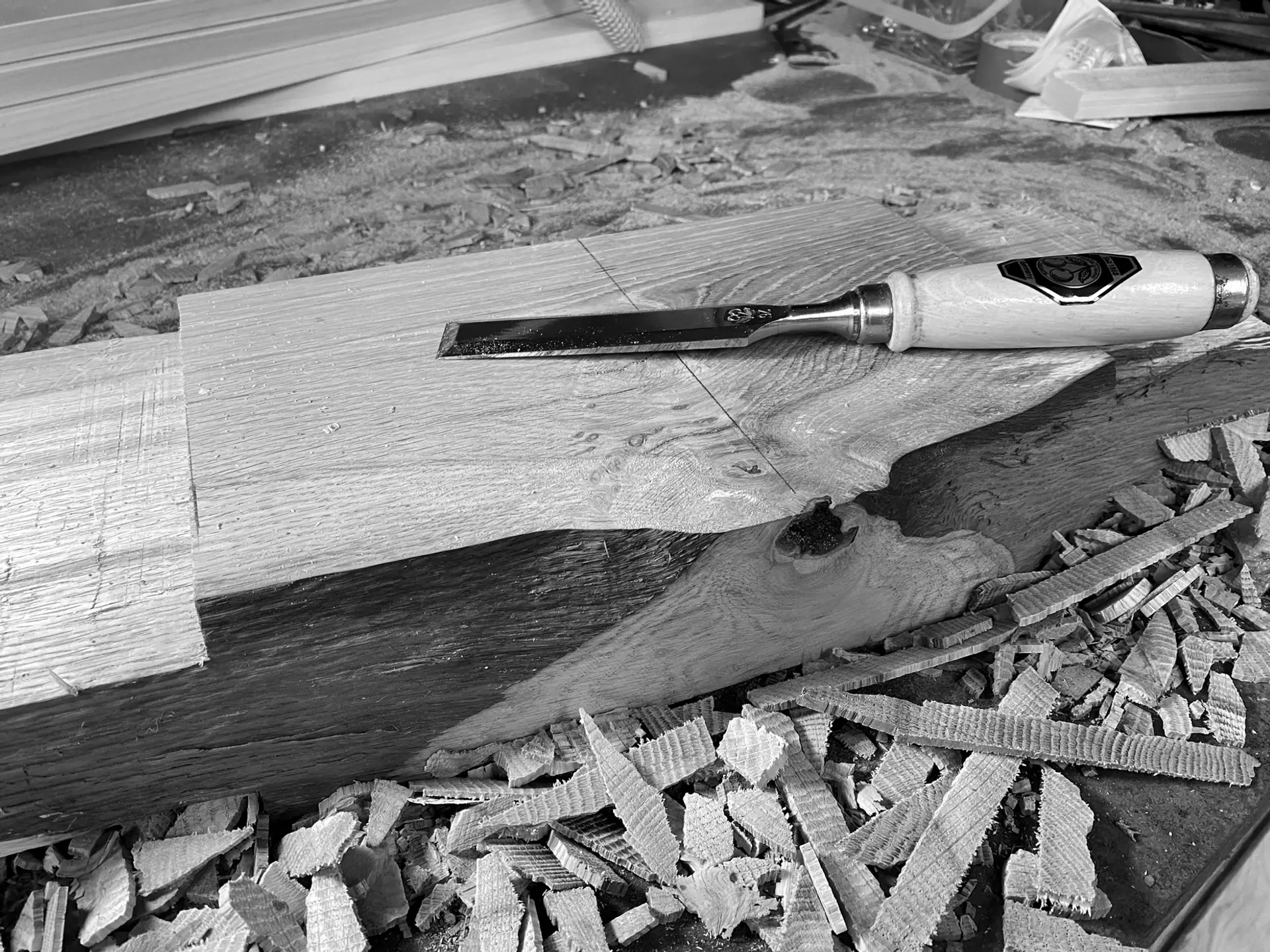 A greyscale photograph showing a plank of wood surrounded by wood shavings and woodchips. A woodwork tool sits on top of the plank, it has a cylindrical wooden handle and a long, flat metal head.