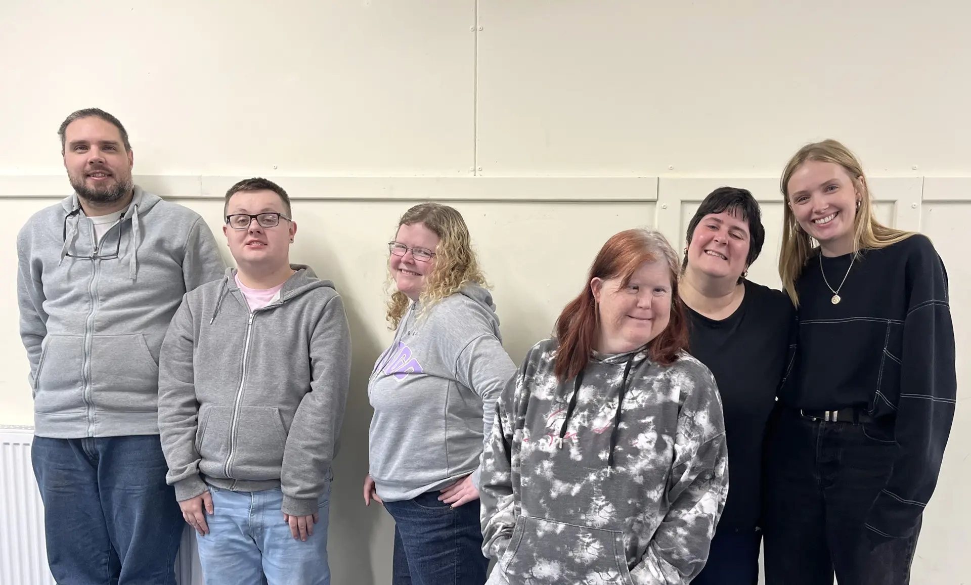 A group photo of the Arbroath pARTicipate group and Lily. They are standing in a line against a cream wall, and are smiling and posing. 