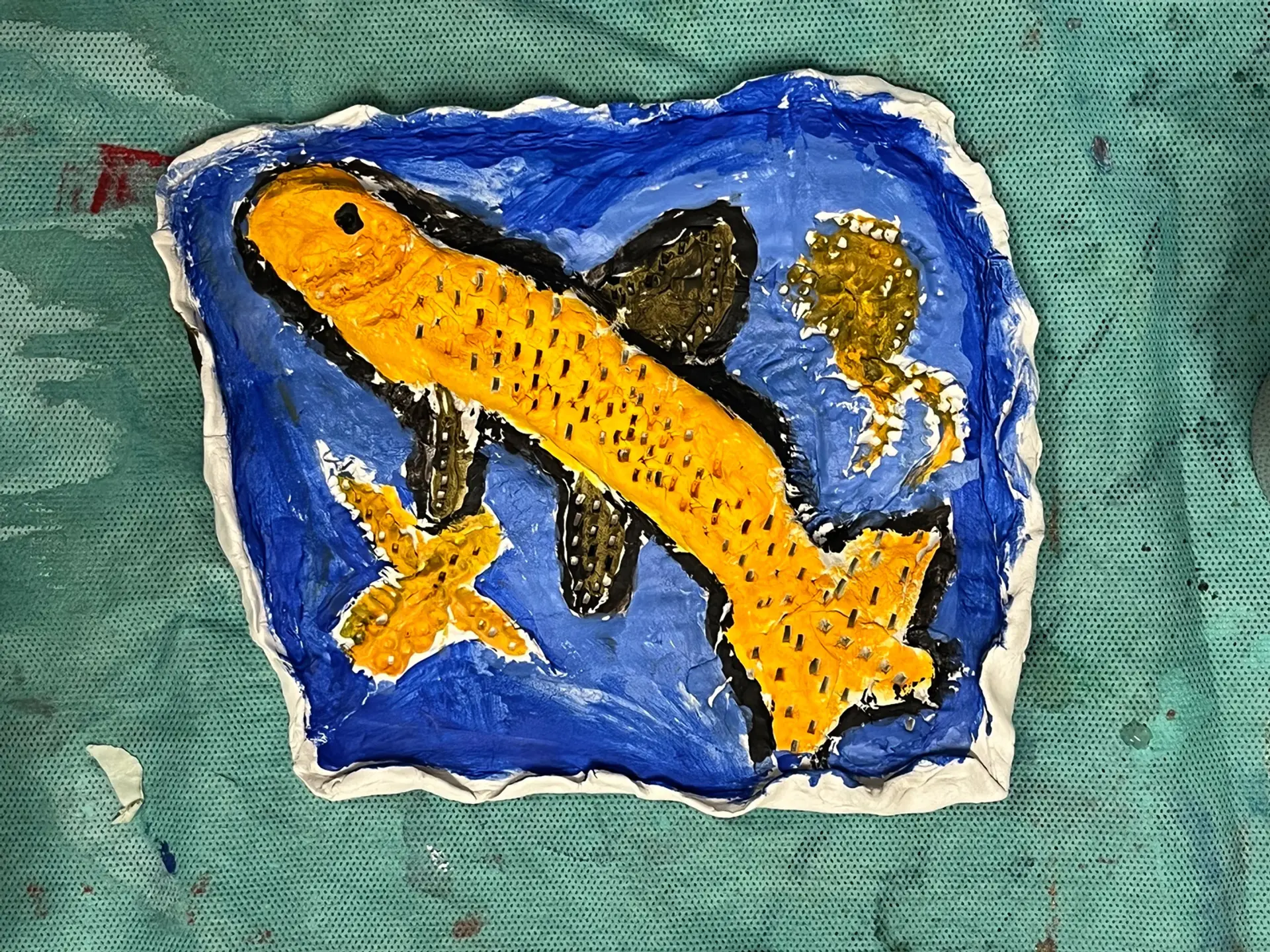 A rectangular tile made from air dried clay, on the tile is a yellow fish with  a blue background and two starfish.