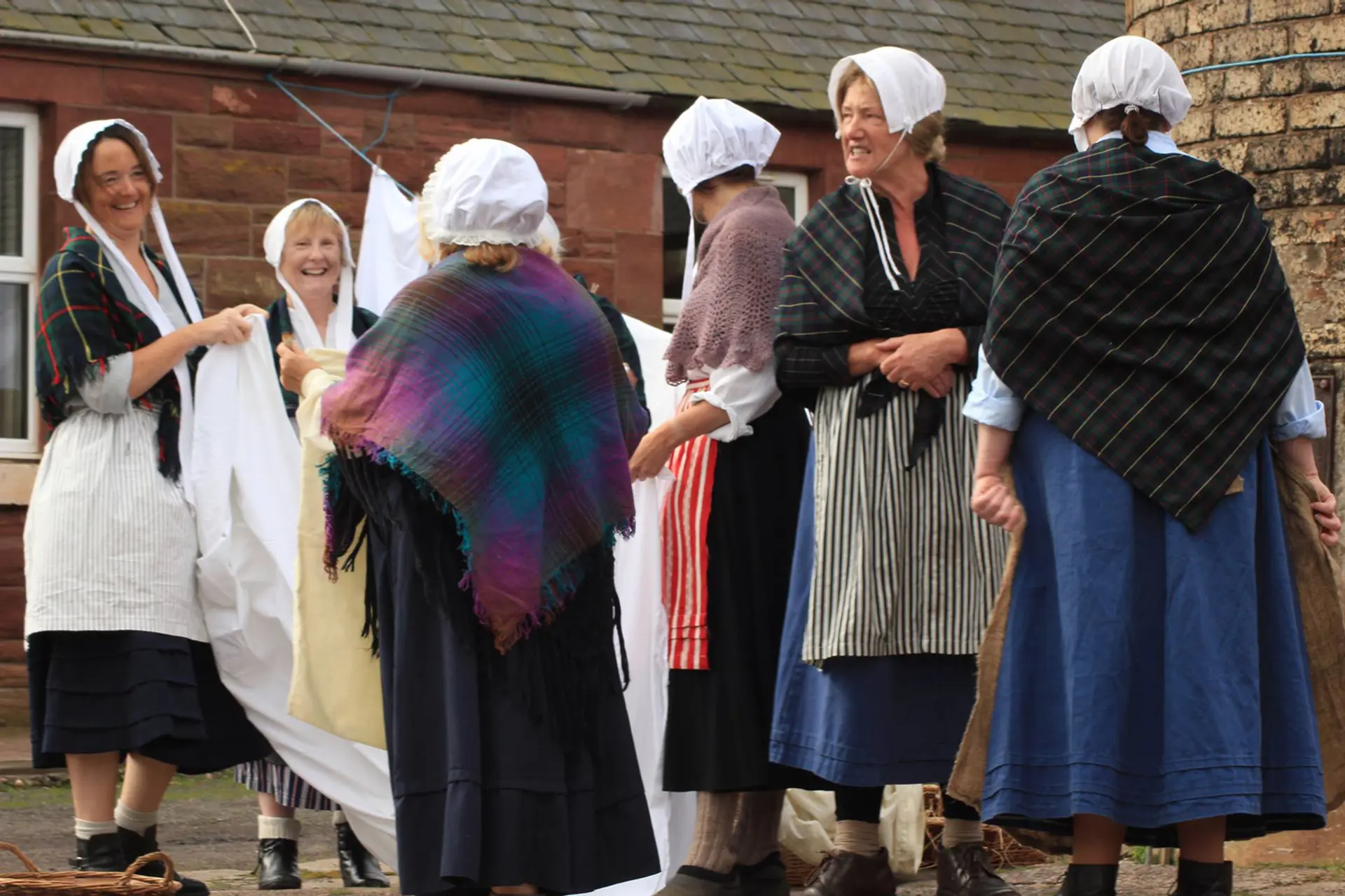 An image of six women dressed up as fishwives, recreating an image from the past. They are laughing and talking. They are wearing long skirts, boots and tartan shawls.