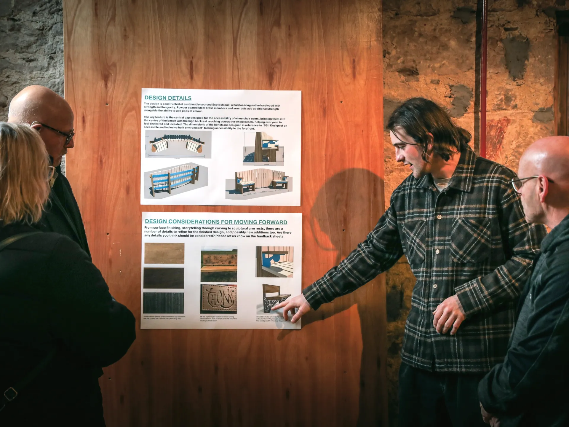 3 people stand looking at posters of bench designs that are mounted into a wooden wall. Another man, in his twenties, points to a part of a design and is talking to the other 3 people.