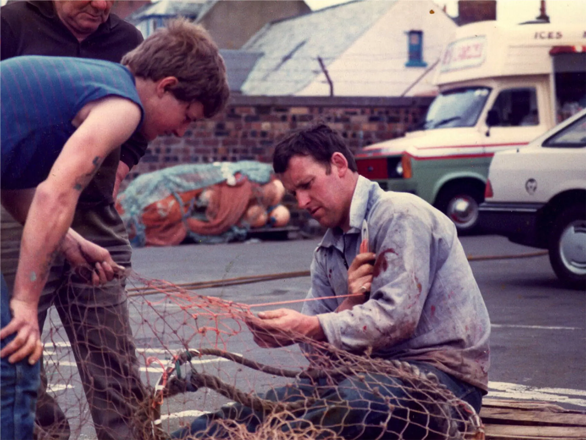 An image from the past, three men are repairing a fishing net in a carpark. 