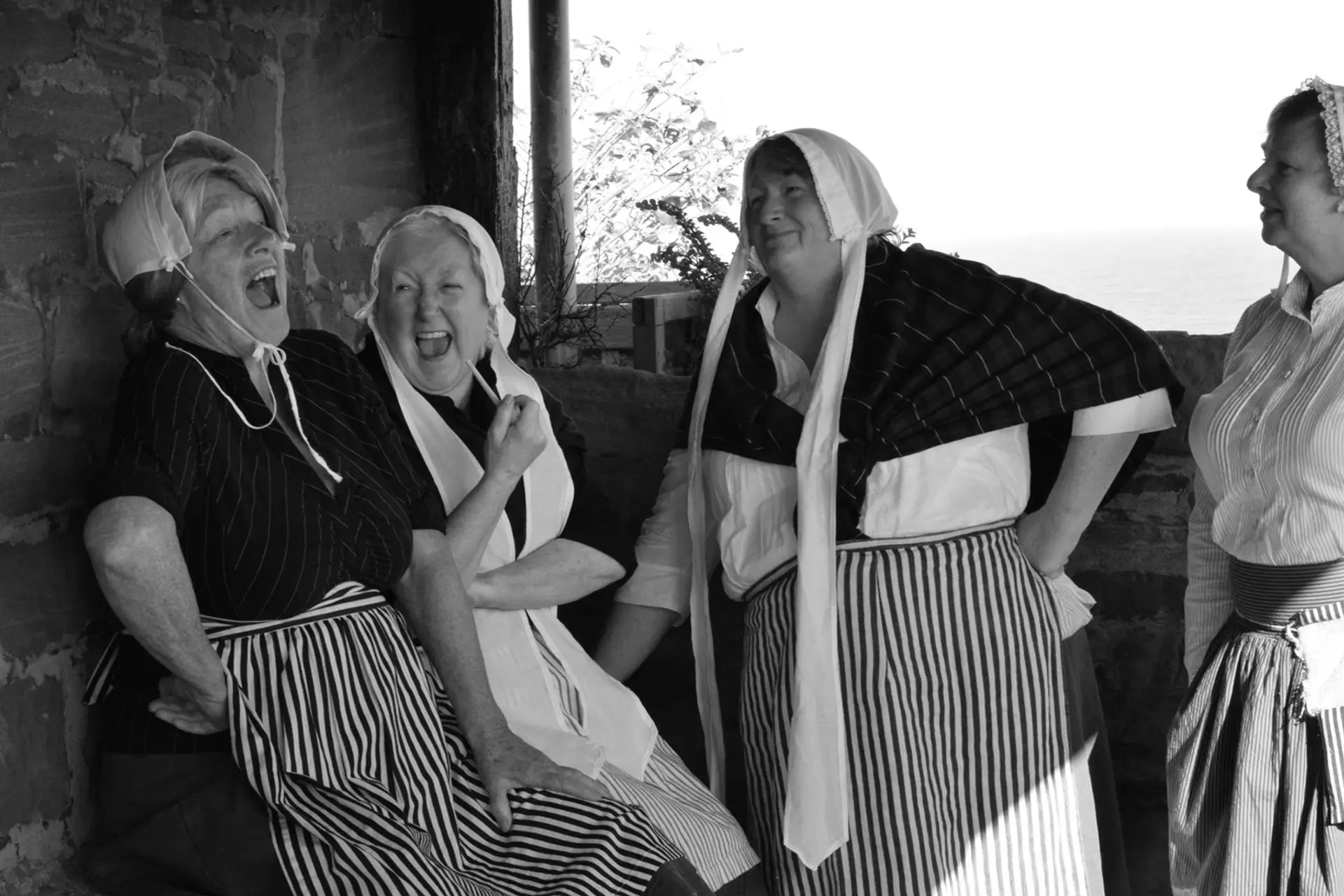 A black and white image of four women dressed up as fishwives, recreating an image from the past. They are laughing and one is smoking a pipe.