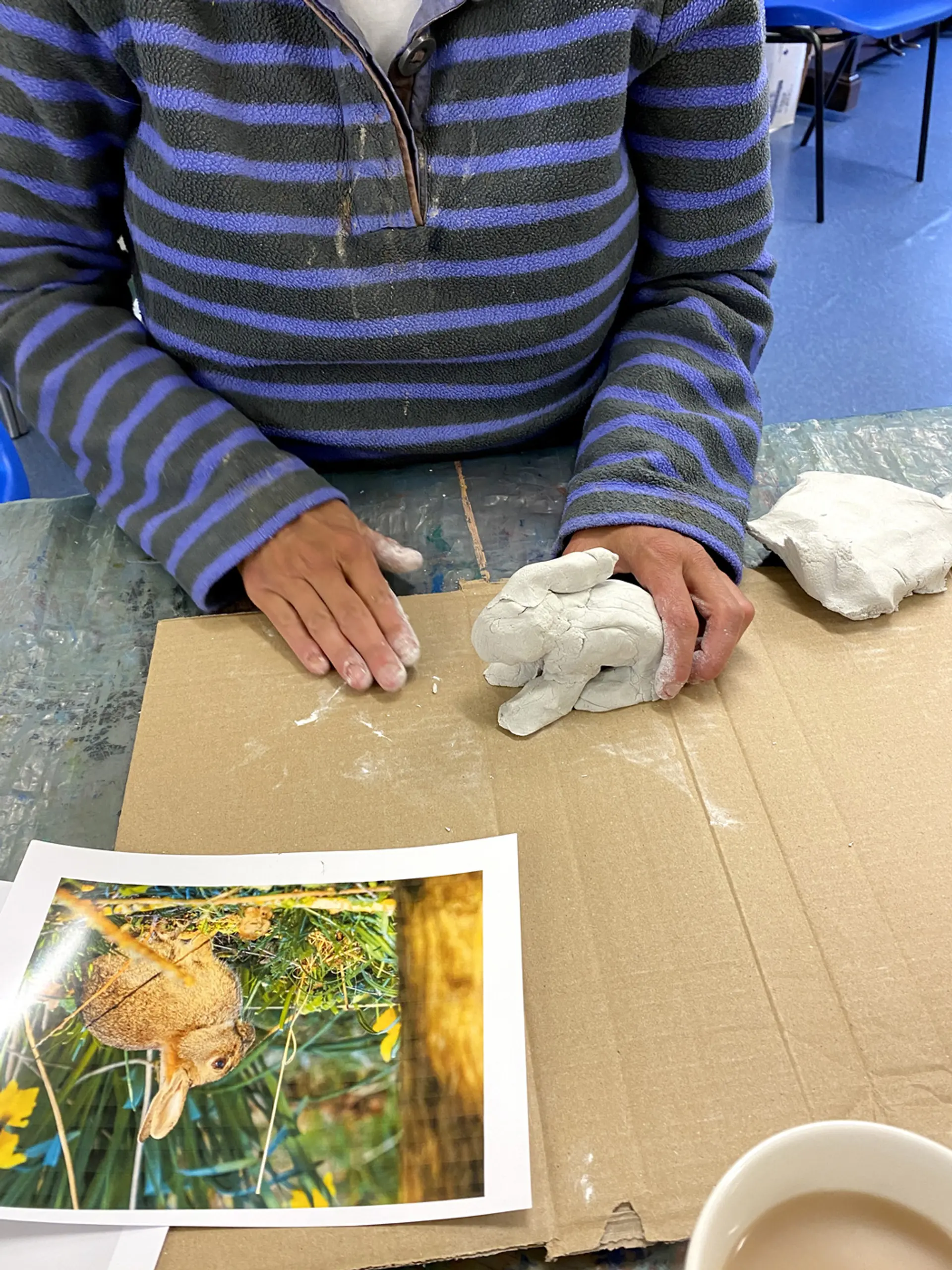 An artist is working with air drying clay on a piece of cardboard. They have an image of a rabbit in front of them and they are sculpting the clay into the shape of a rabbit. 