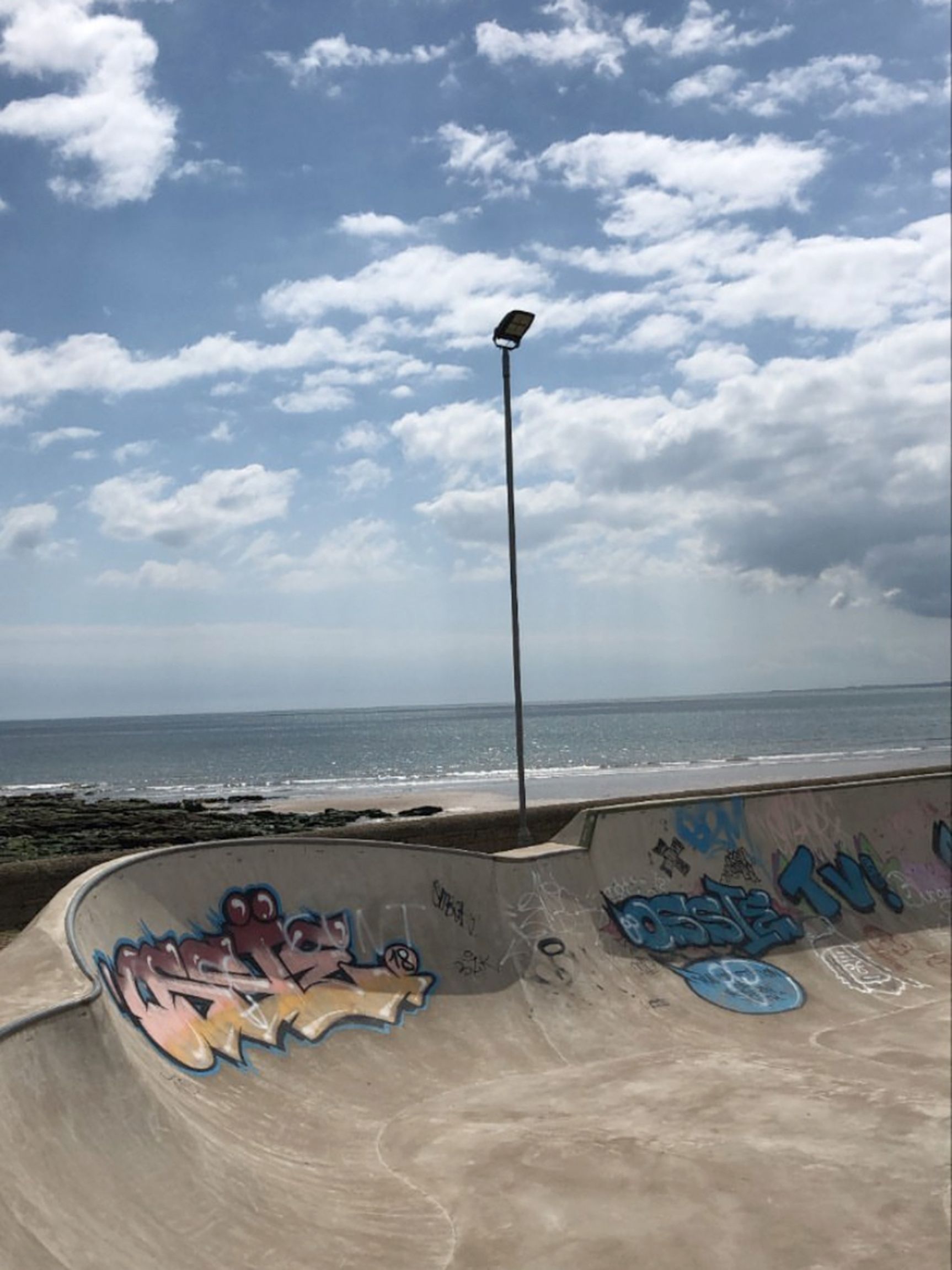 In the foreground is one of the concrete ramps at the Carnoustie skatepark, overlooking the rocks, sandy beach and blue waves of the North Sea.