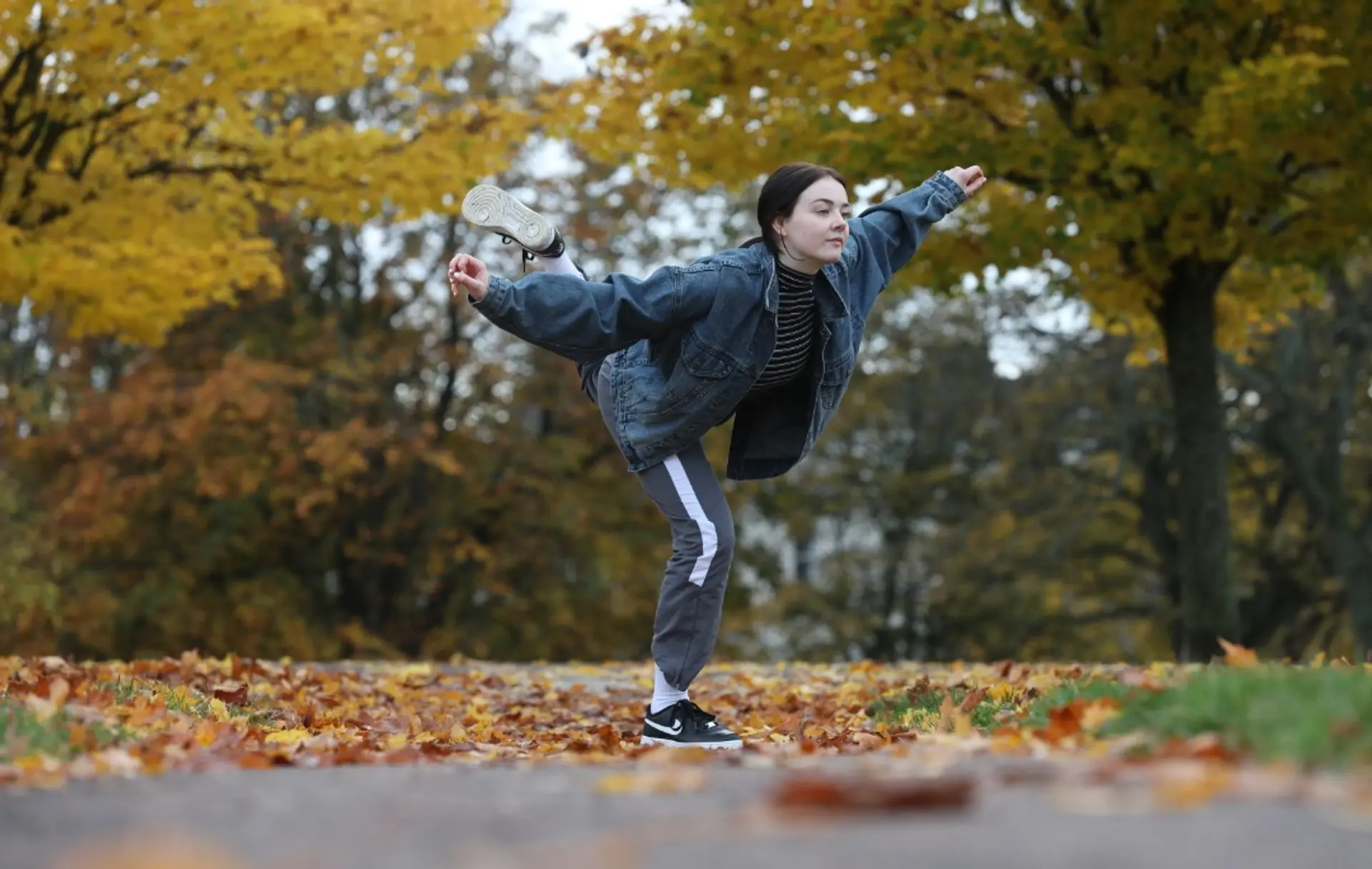 An image of a dancer posing on one bent leg, their other leg is extended behind them. Their arms are extended out from their sides. They are pictured in an autumn park with leaves on the ground and trees with yellow leaves in the background. 