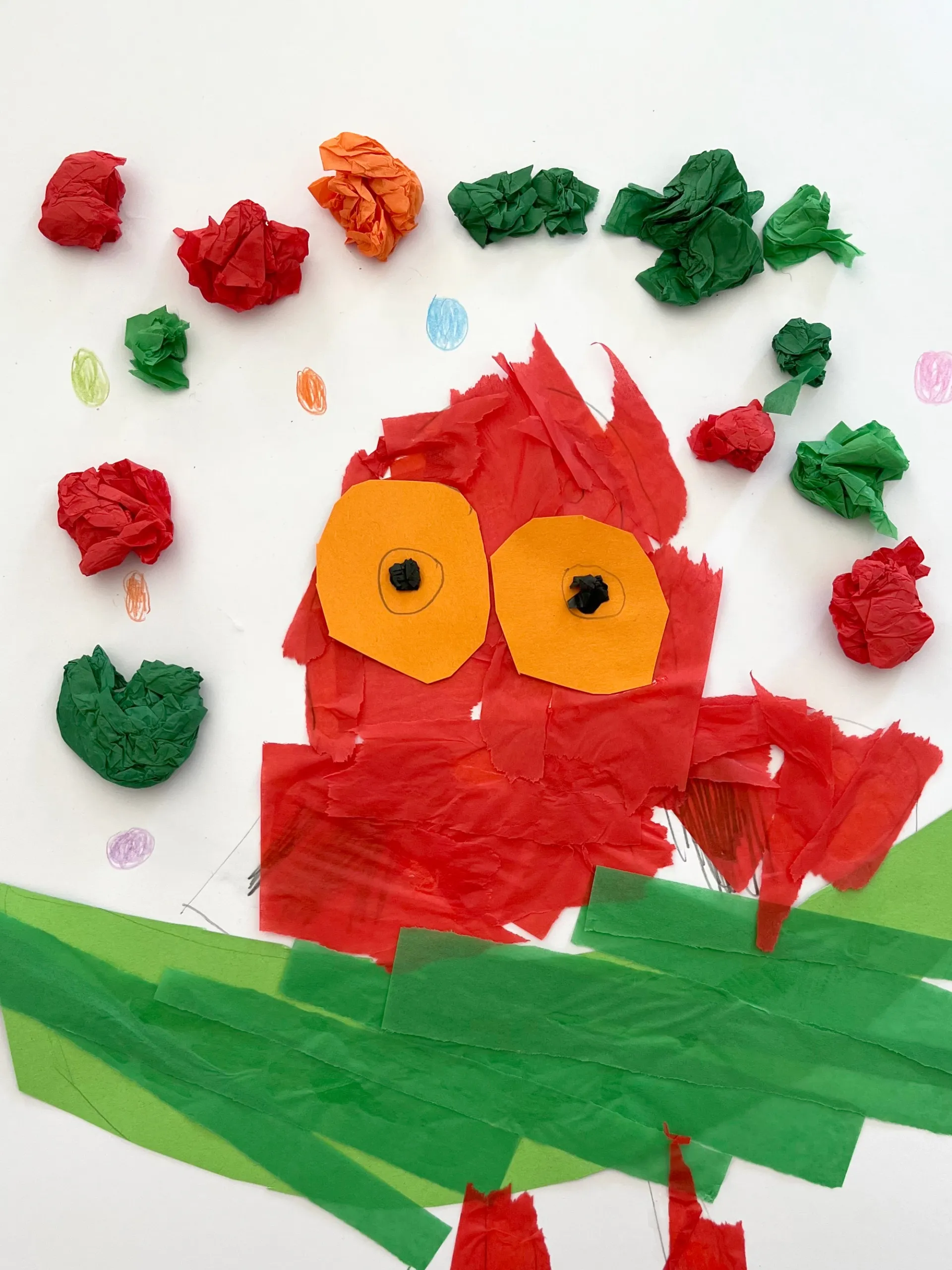 A collage of an owl made from red tissue paper. The owl is sitting on a green branch and has big orange eyes. There are scrunched up pieces of green, red and orange tissue paper in the sky around them.