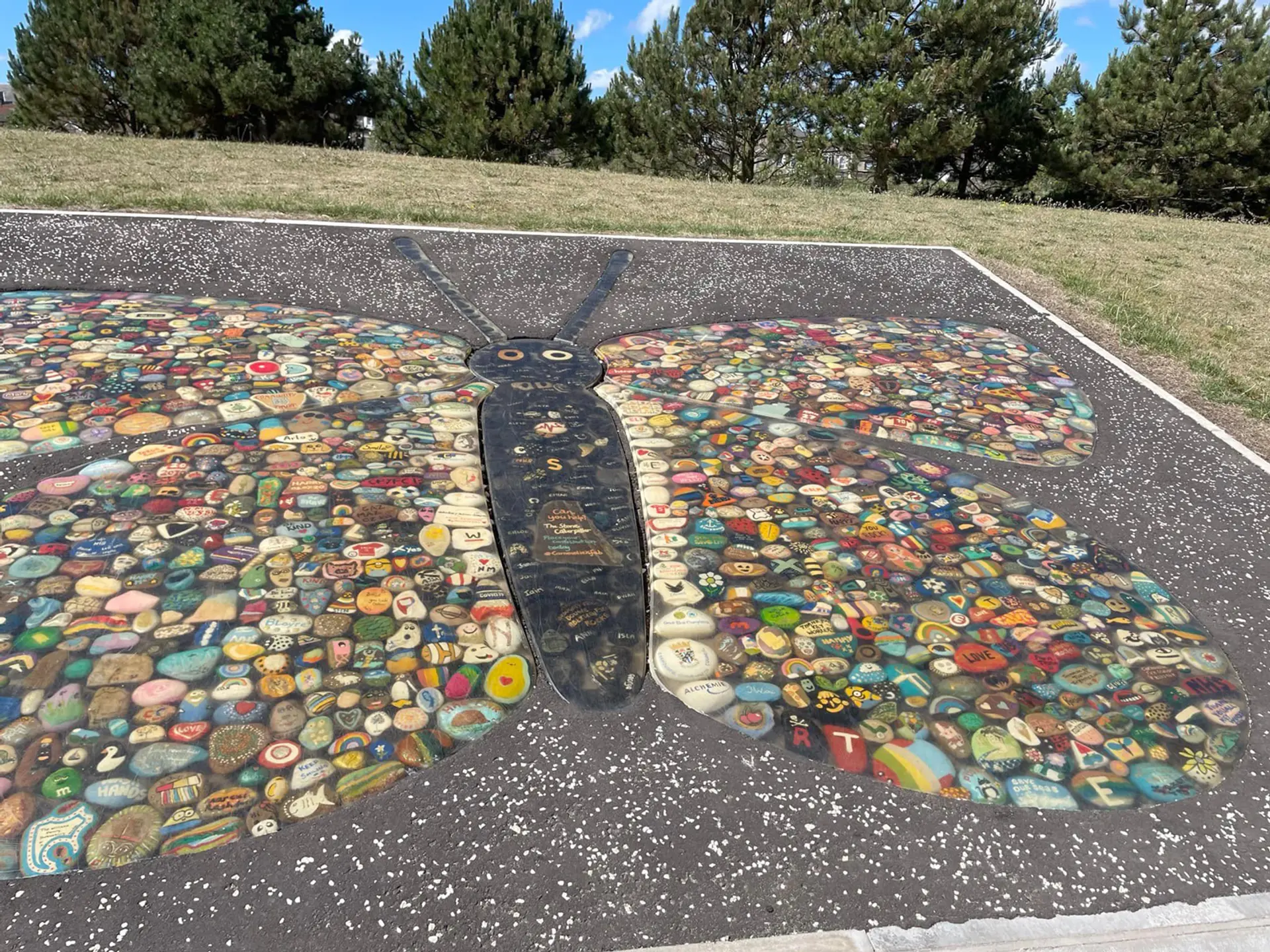 A large butterfly has been created in a tarmac surround using pebbles painted by the local community during the pandemic. The wings of the butterfly are colourful and the body, head and antennae are coloured black. Around the tarmac is green grass and trees.