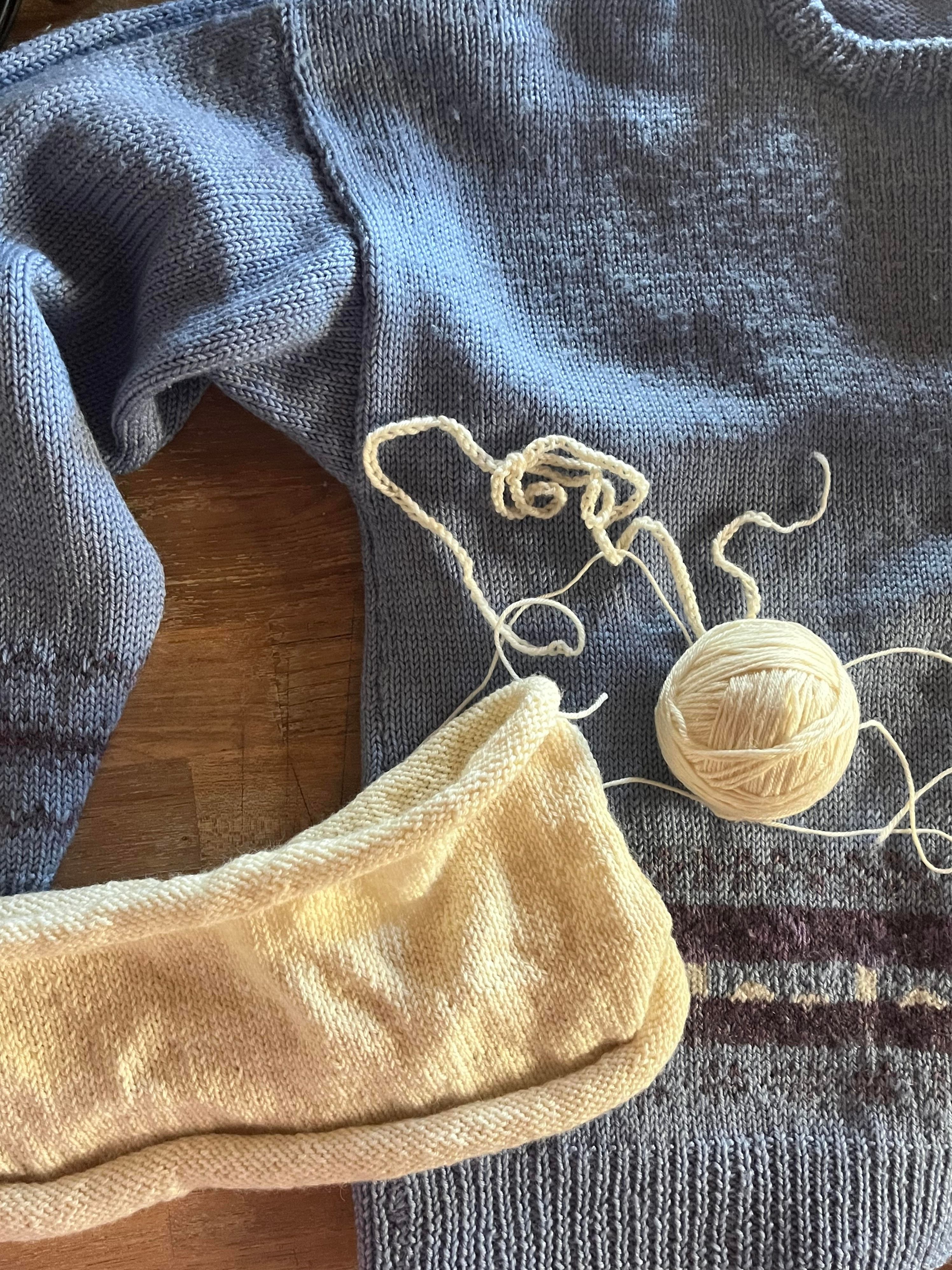 A cropped image of a blue hand knitted jumper with purple detailing is laid flat on a wooden table. A half finished cream jumper is placed over the top, being compared for size.