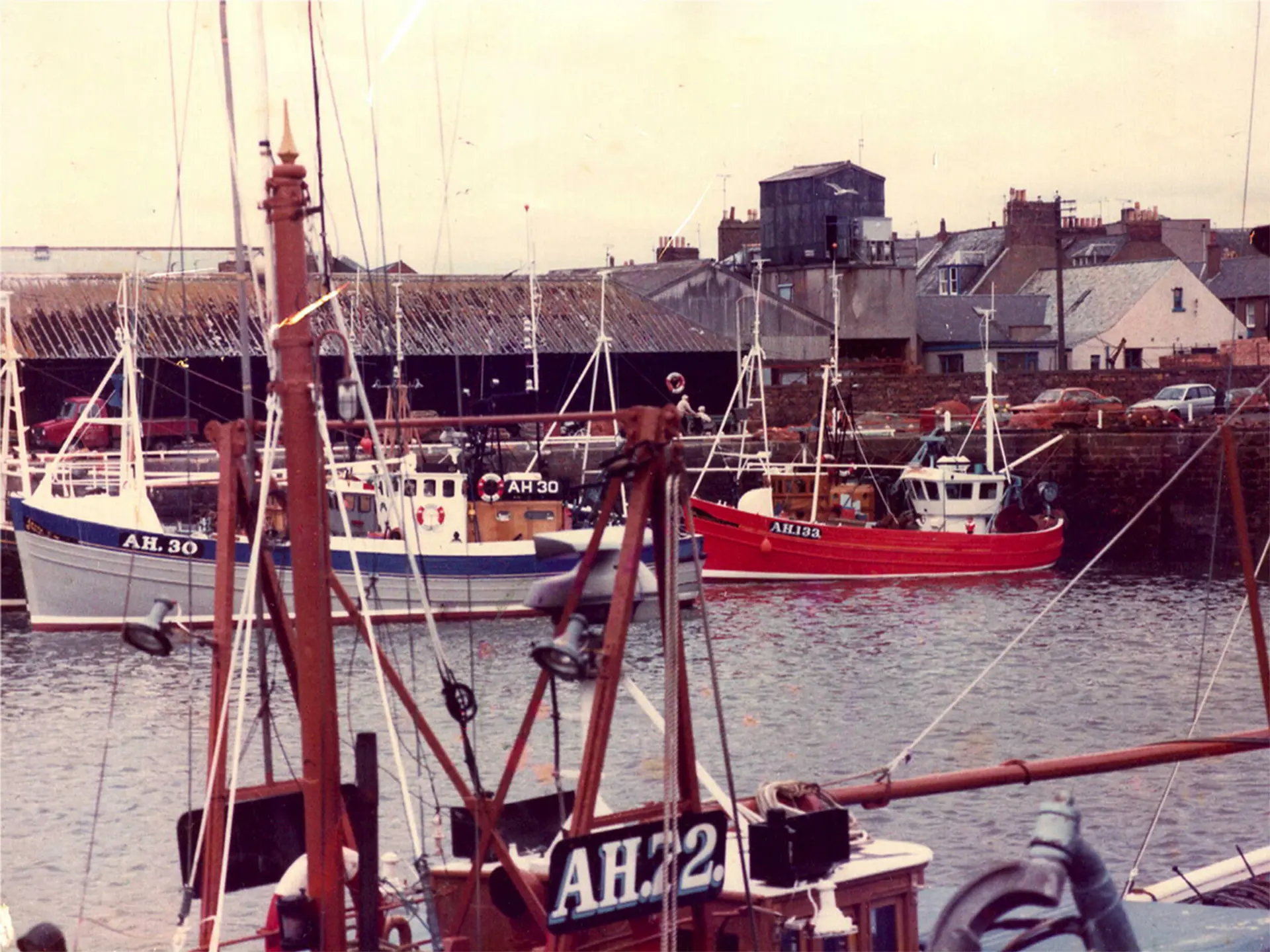 An image of Arbroath harbour from the past. We are looking through the mast of a boat, and can clearly see two fishing boats, one red and one white and blue. 