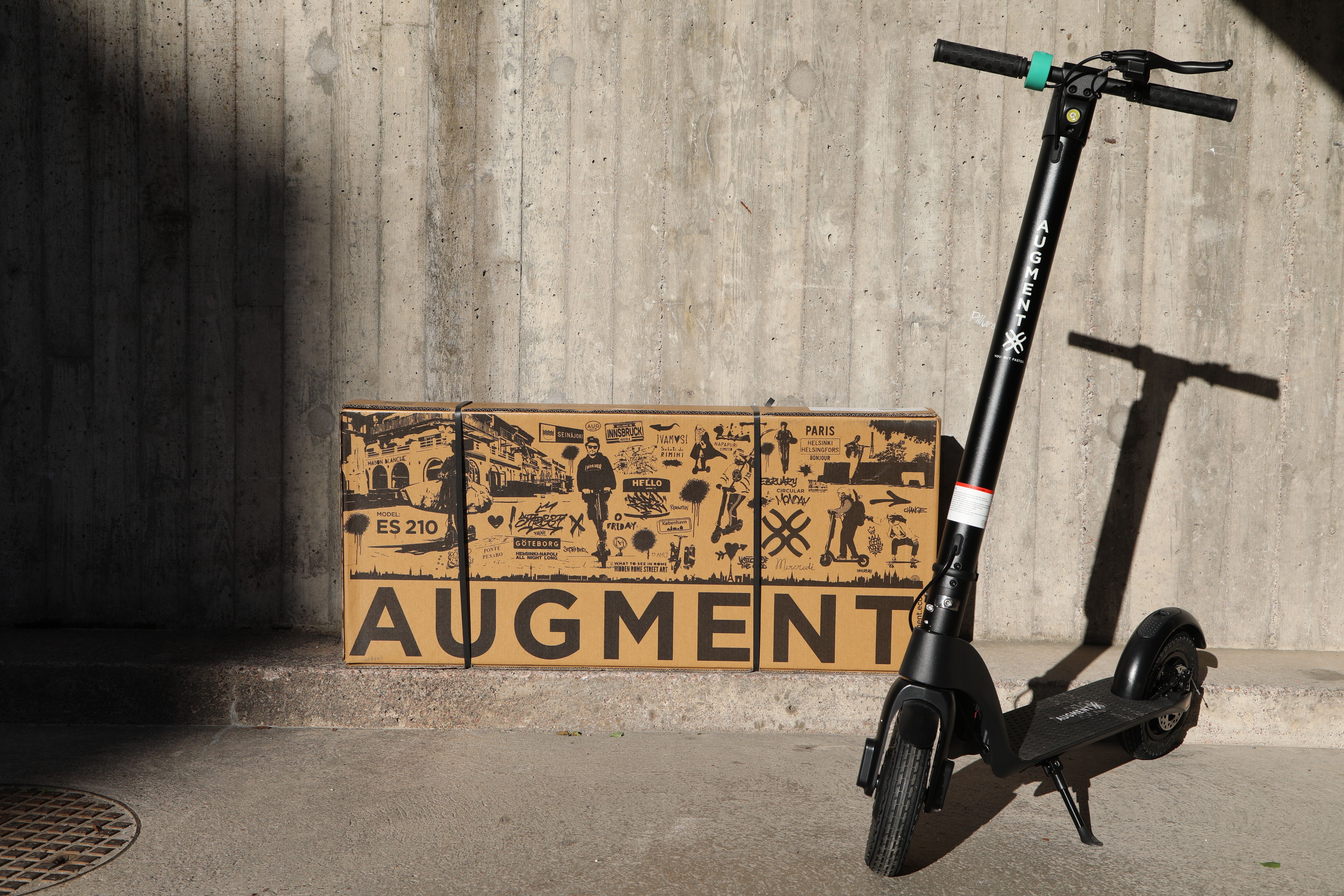 Vista frontale di un e-scooter Augment davanti al muro, su cui è appoggiato il pacco postale Augment