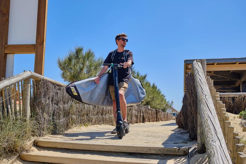 Un homme avec un scooter électrique Augment sur une route de plage sablonneuse avec une planche de surf.