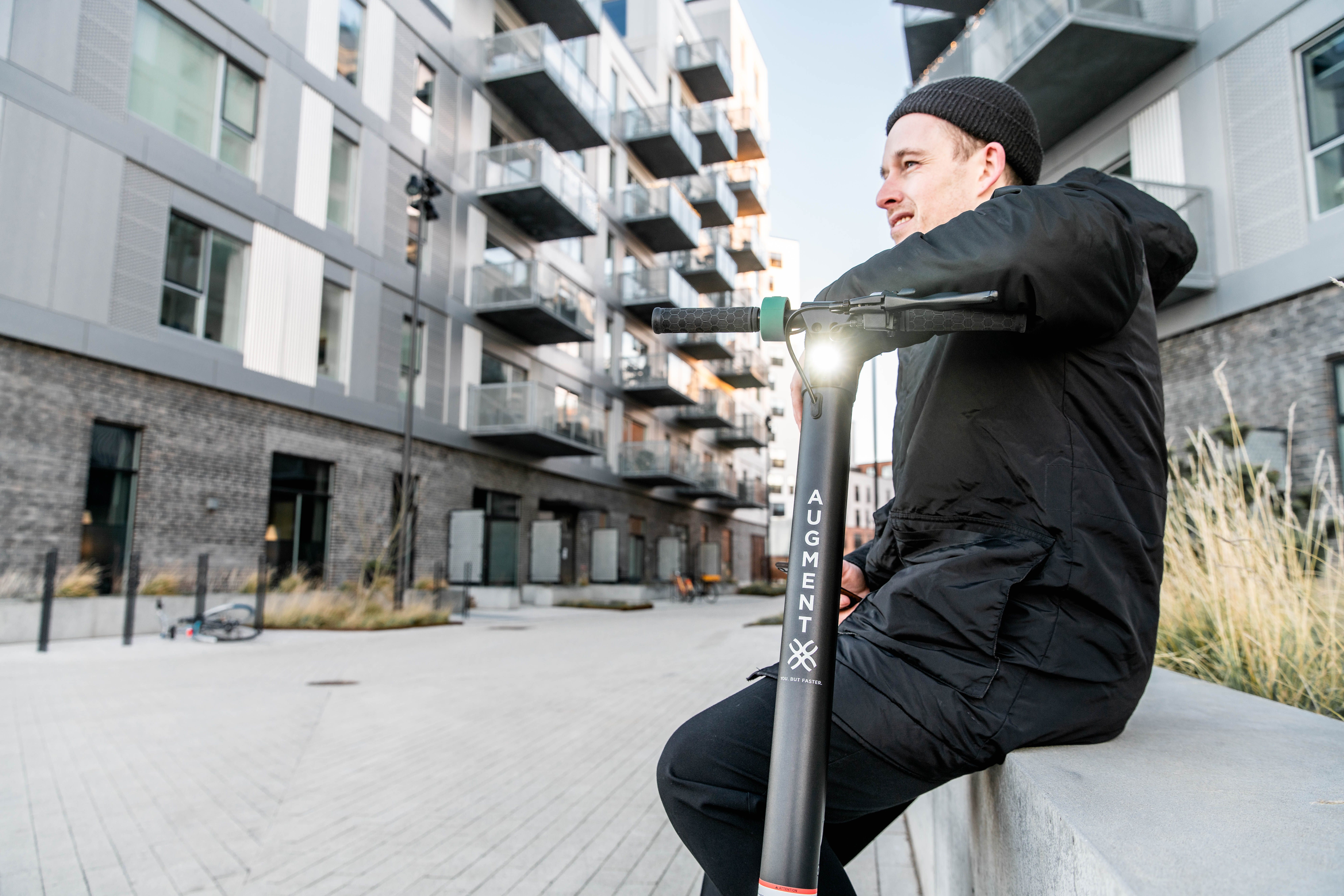 En mand sidder på en væg i midten af to boligblokke med en Augment e-scooter