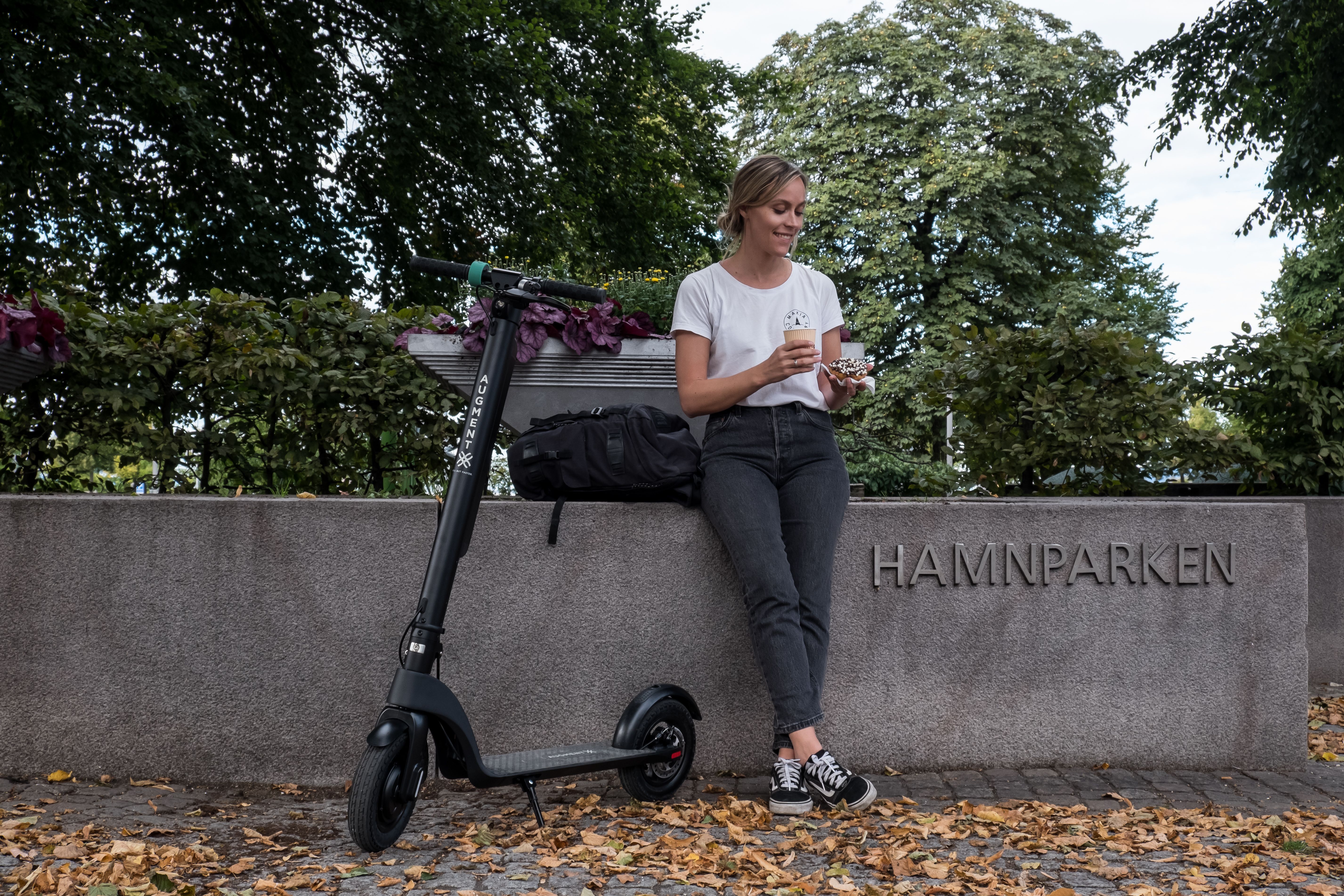 En kvinde holder kaffepause efter at have kørt på en Augment e-scooter ved siden af en bypark