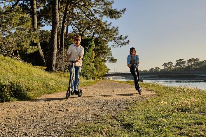Two people riding e-scooter by a river