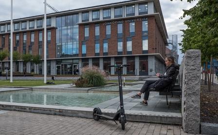 En kvinna sitter på en parkbänk framför Jönköpings universitet bredvid en Augment-elrullator.