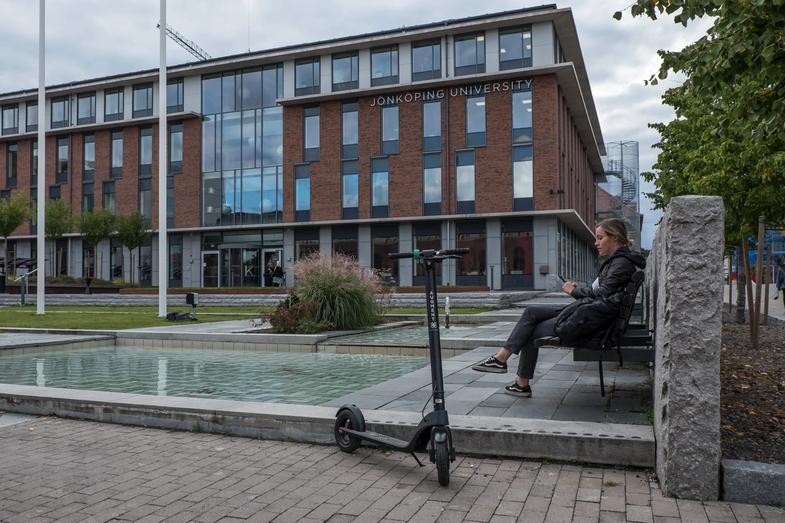 En kvinna sitter på en parkbänk framför Jönköpings universitet bredvid en Augment-elrullator.
