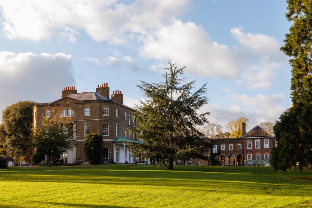 Scenic view of Belmont, Mill Hill Prep, showing the historic building surrounded by lush greenery and tall trees under a partly cloudy sky, highlighting the peaceful campus grounds.