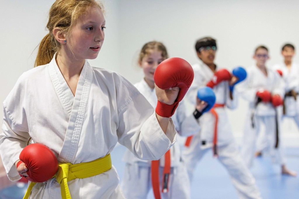 Students at Belmont, Mill Hill Prep, practicing karate in a group, focused and wearing traditional white uniforms with coloured belts and protective gloves during a martial arts session.