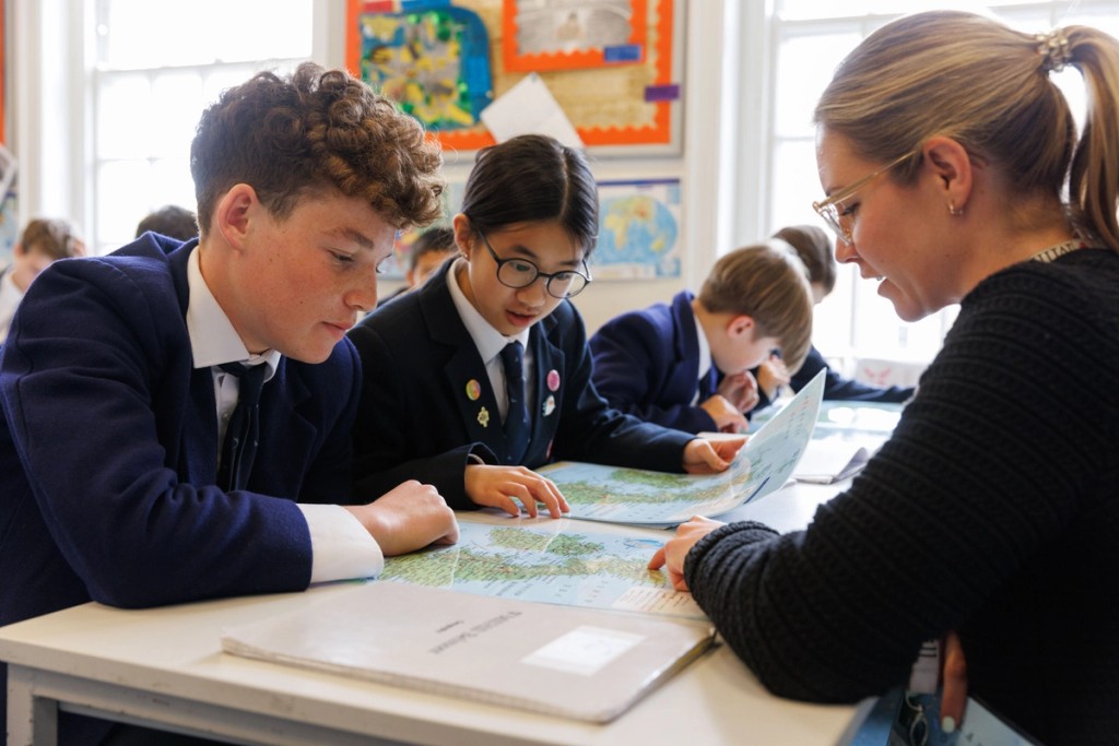 Students at Belmont, Mill Hill Prep, working together with a teacher on a geography activity, engaged in reading maps and discussing in a classroom environment filled with educational materials.