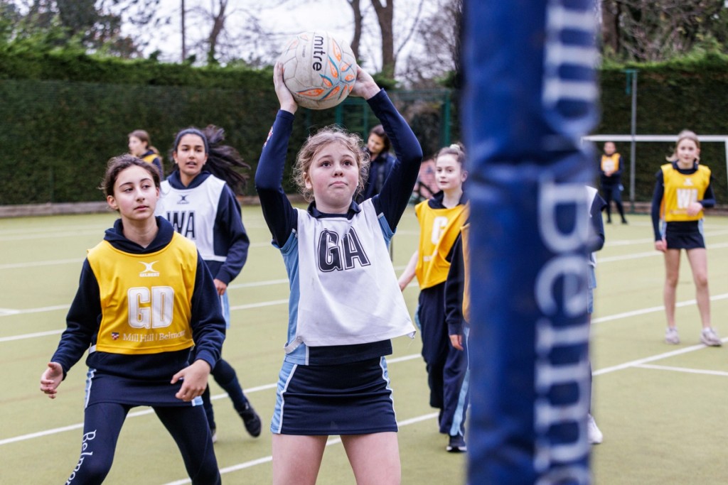 Girls at Belmont, Mill Hill Prep, engaged in a competitive netball match, with a player in the Goal Attack (GA) position preparing to take a shot, while teammates and opponents focus on the game around her.