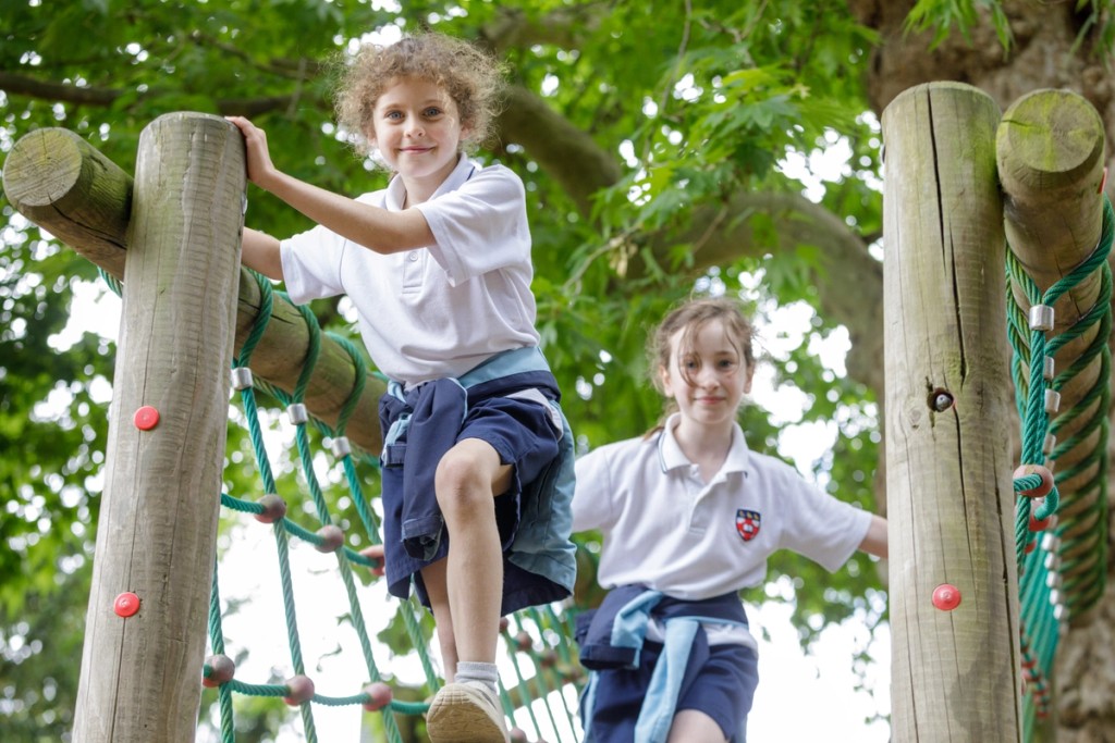 Two students enjoy climbing on a wooden playground structure, smiling as they navigate an outdoor adventure course, surrounded by greenery.