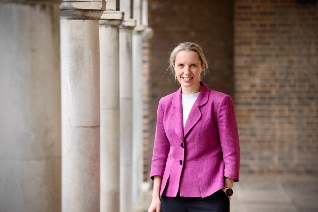 Portrait of the Head of Belmont, Mill Hill Prep, standing outdoors in a columned walkway, smiling and wearing a pink blazer, creating a professional and approachable image.