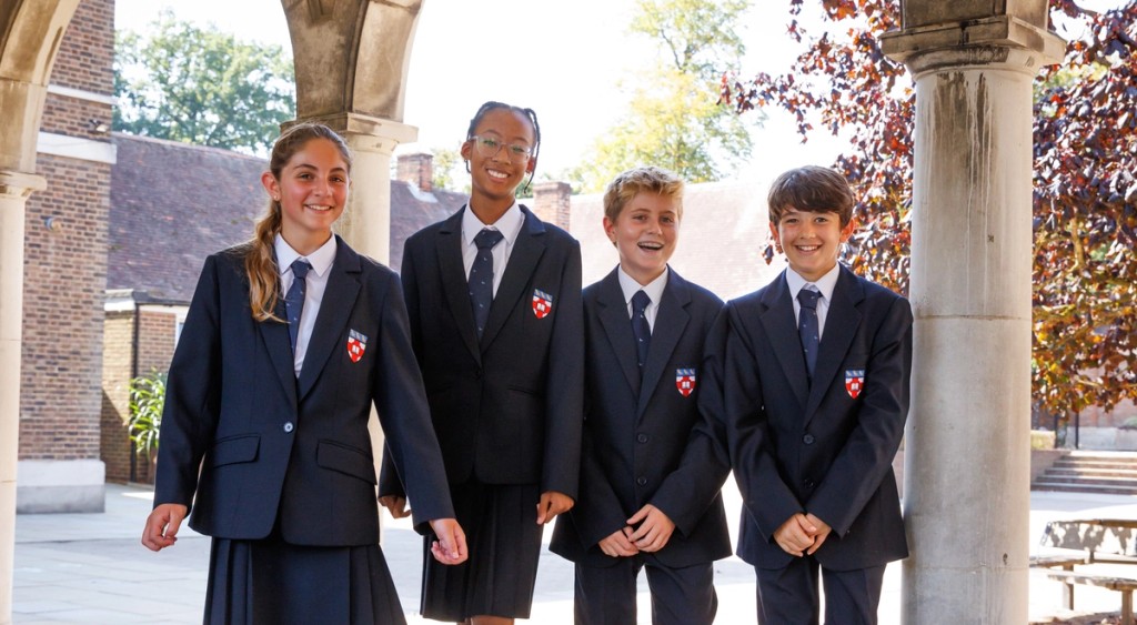 A group of four students walks together under an archway, smiling and chatting in their formal uniforms, enjoying a sunny day at school.