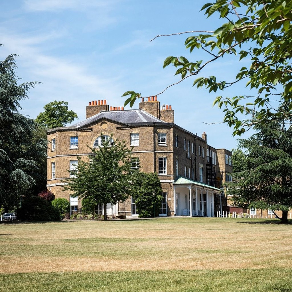 Exterior view of Belmont, Mill Hill Prep, featuring a historic brick building surrounded by greenery, with a large lawn in the foreground under a clear blue sky.