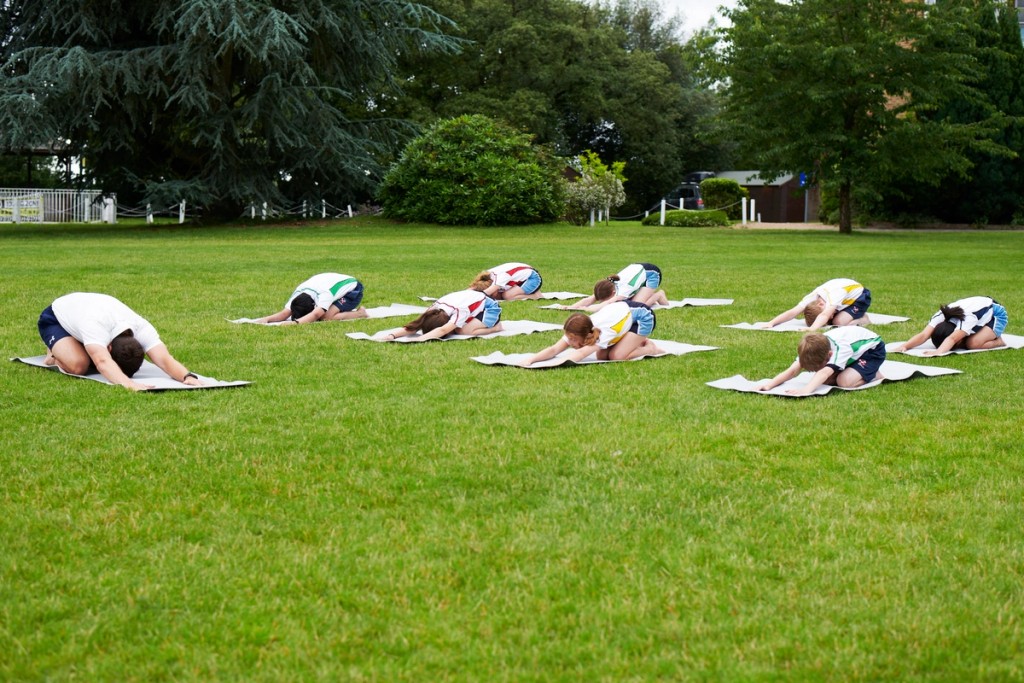 Students at Belmont, Mill Hill Prep, practicing yoga or stretching exercises outdoors on a grassy field, each on their own mat, surrounded by greenery and trees in a peaceful setting.