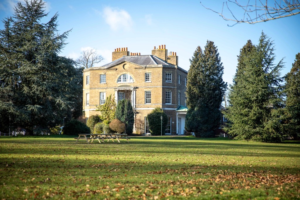 Exterior view of Belmont, Mill Hill Prep, featuring a historic brick building surrounded by greenery, with a large lawn in the foreground under a clear blue sky.