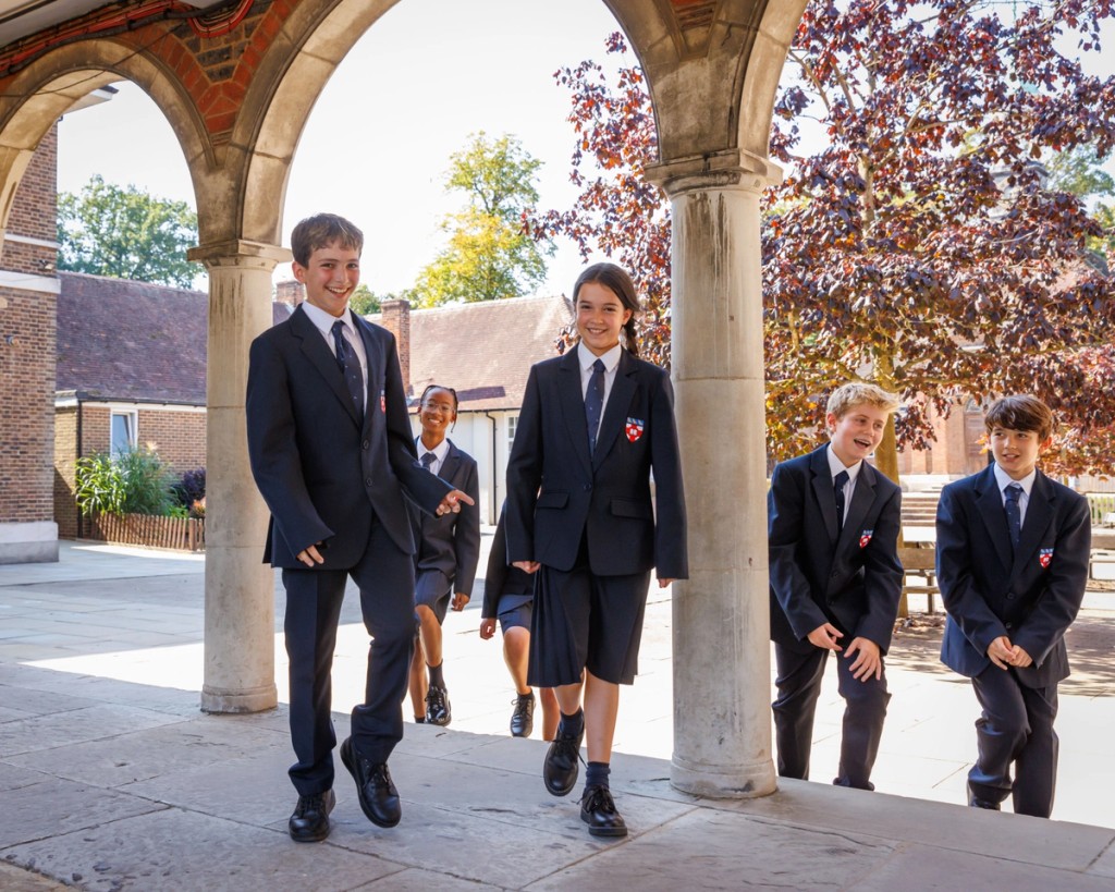 A group of four students walks together under an archway, smiling and chatting in their formal uniforms, enjoying a sunny day at school.