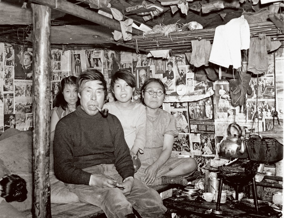 Ill. 2. Indenfor hos Nikolaj og Sofie Maqe og deres yngste børn på bopladsen Akerninnaq, ca. 1955. Avissiderne på væggen fungerer som tapet. Efter sigende var dette hjem særligt overdådigt i så henseende. Man ser både den tændte spæklampe og tørreribben på billedet.