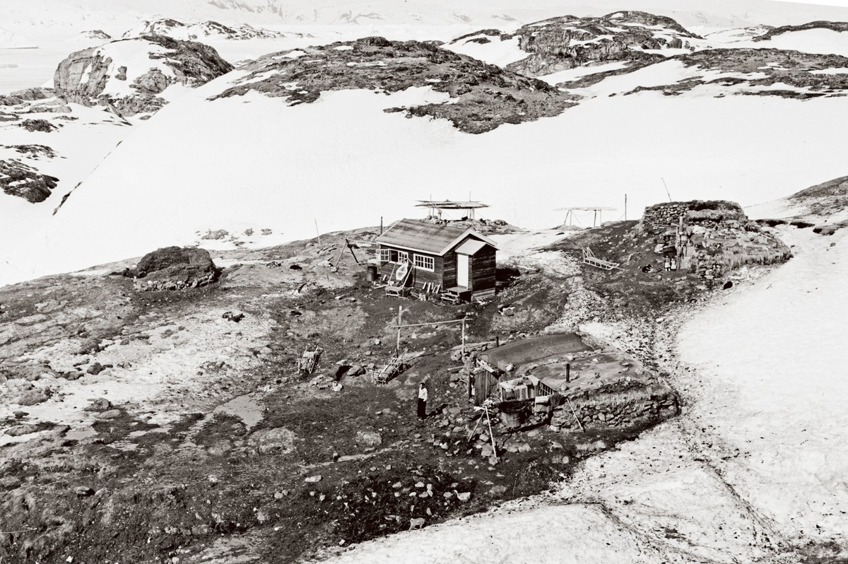 Ill. 5. Ude på de små steder boede mange stadig i tørvemurshuse i 1950’erne. Her ses en ruin til venstre og til højre to fungerende huse. Desuden er der bygget et nyt bræddehus. Bag husene ses kajakker, der ligger på stativer, så hundene ikke kan komme til at gnave i dem. Den nu affolkede boplads hed Ikkatteq.