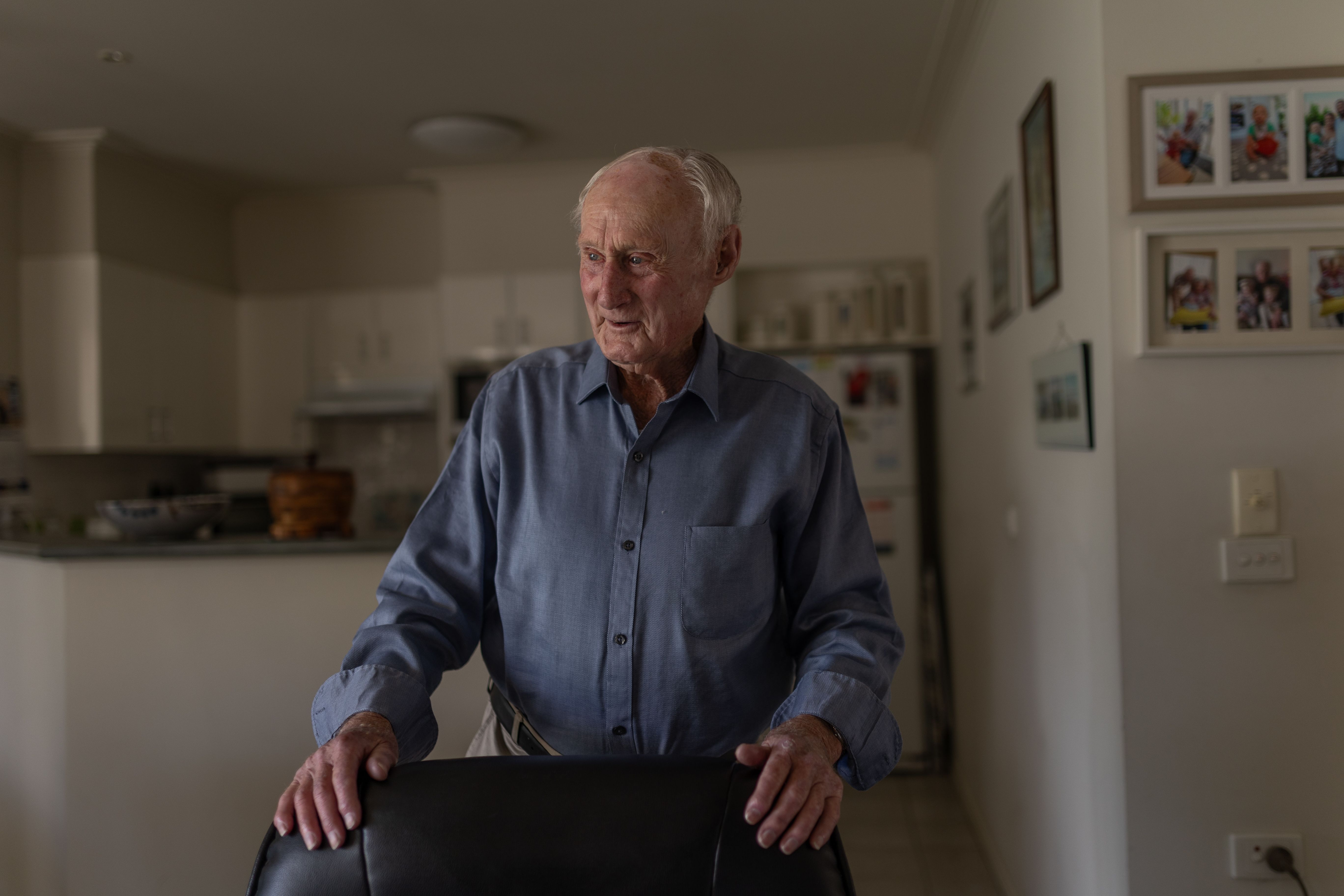 Older man in his 80s standing in his living room, with his hands resting on the back of his armchair, smiling and looking away