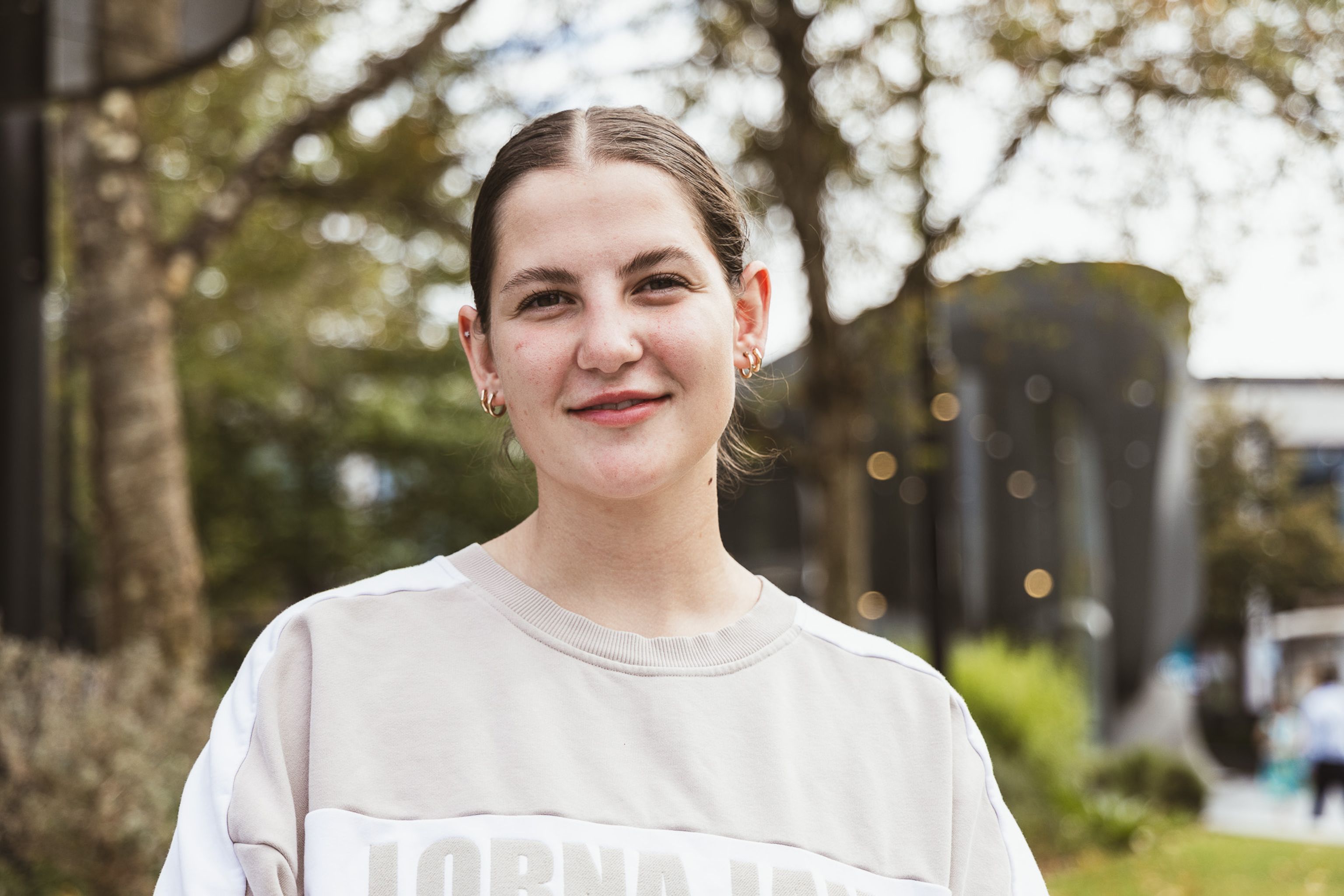 Abbey, a young woman with light skin and dark brown hair stands outdoors, looking forward with a gentle smile.