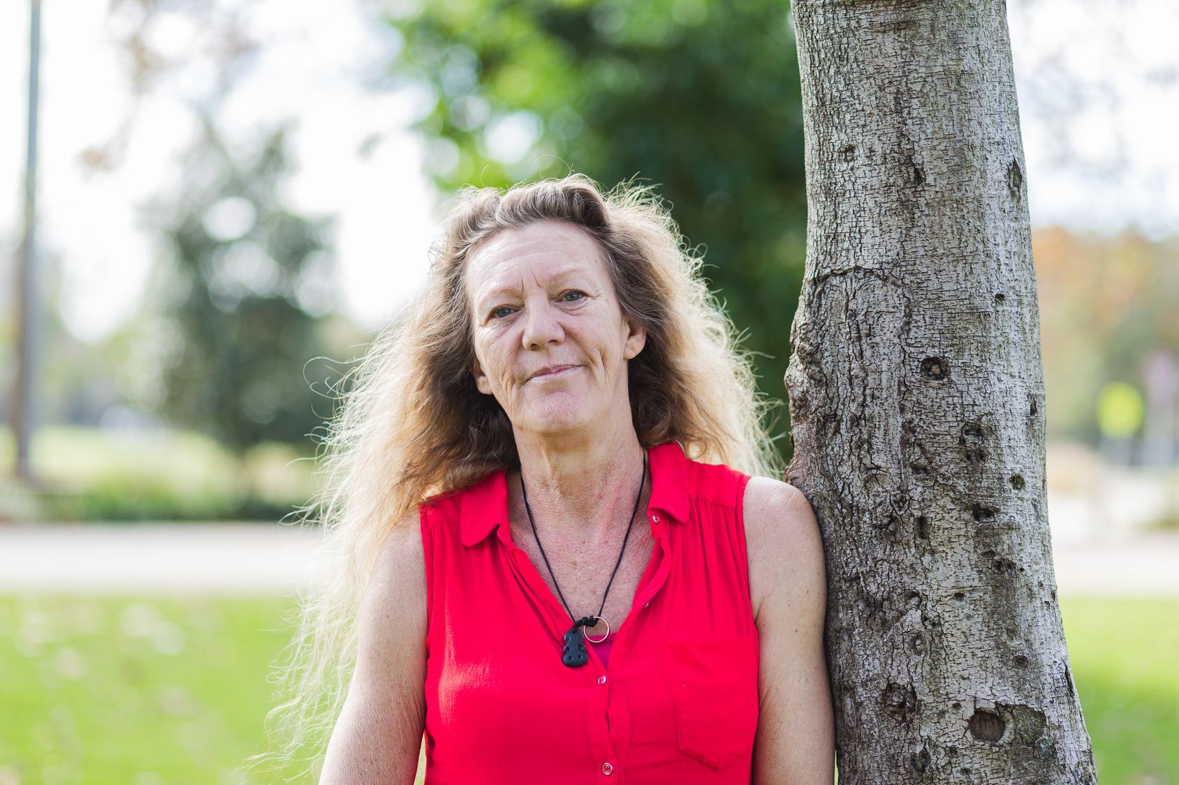 Woman in a bright red sleeveless shirt standing outdoors next to a tree, with green foliage and a park-like background.