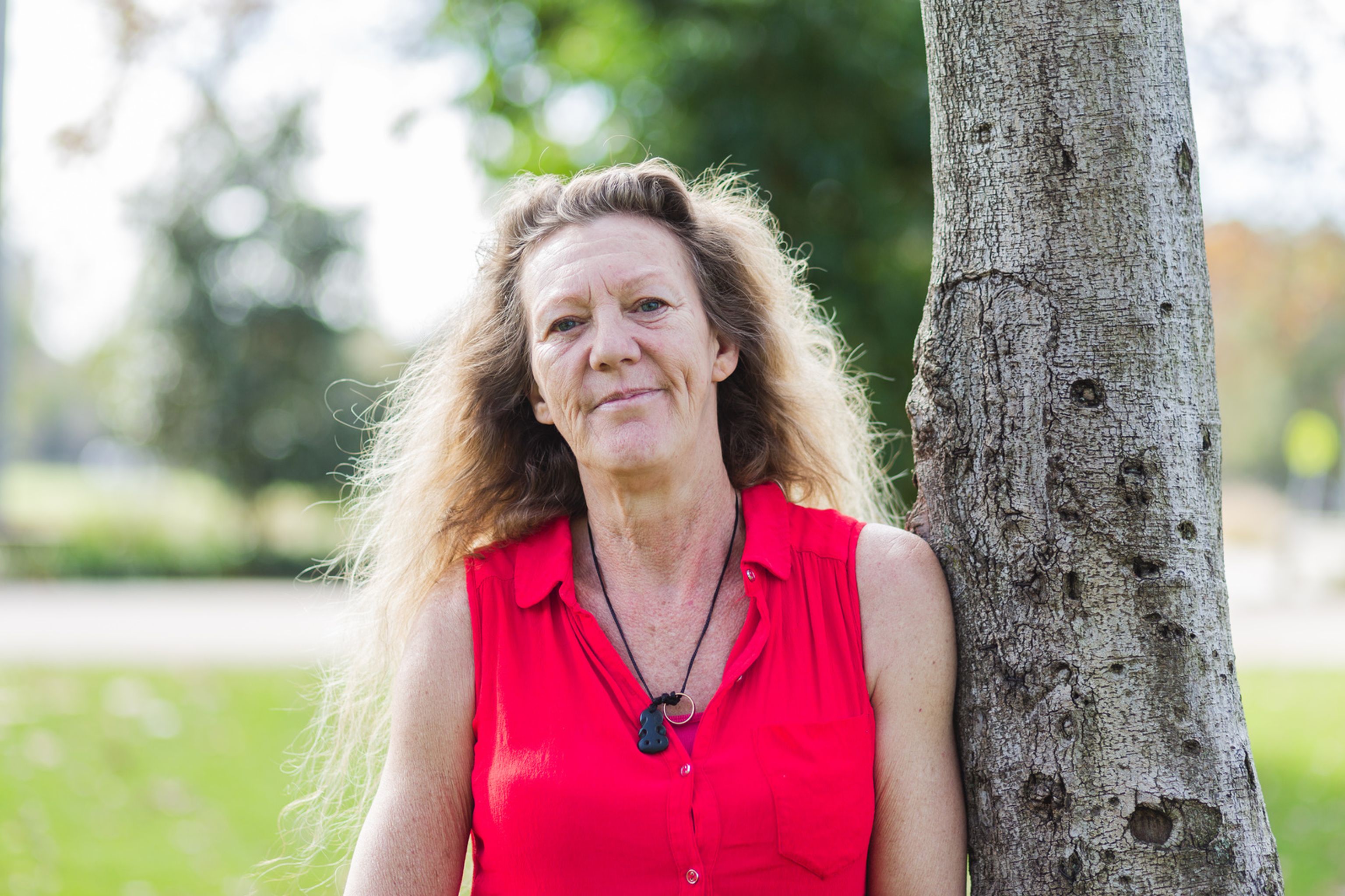 Woman in a bright red sleeveless shirt standing outdoors next to a tree, with green foliage and a park-like background.