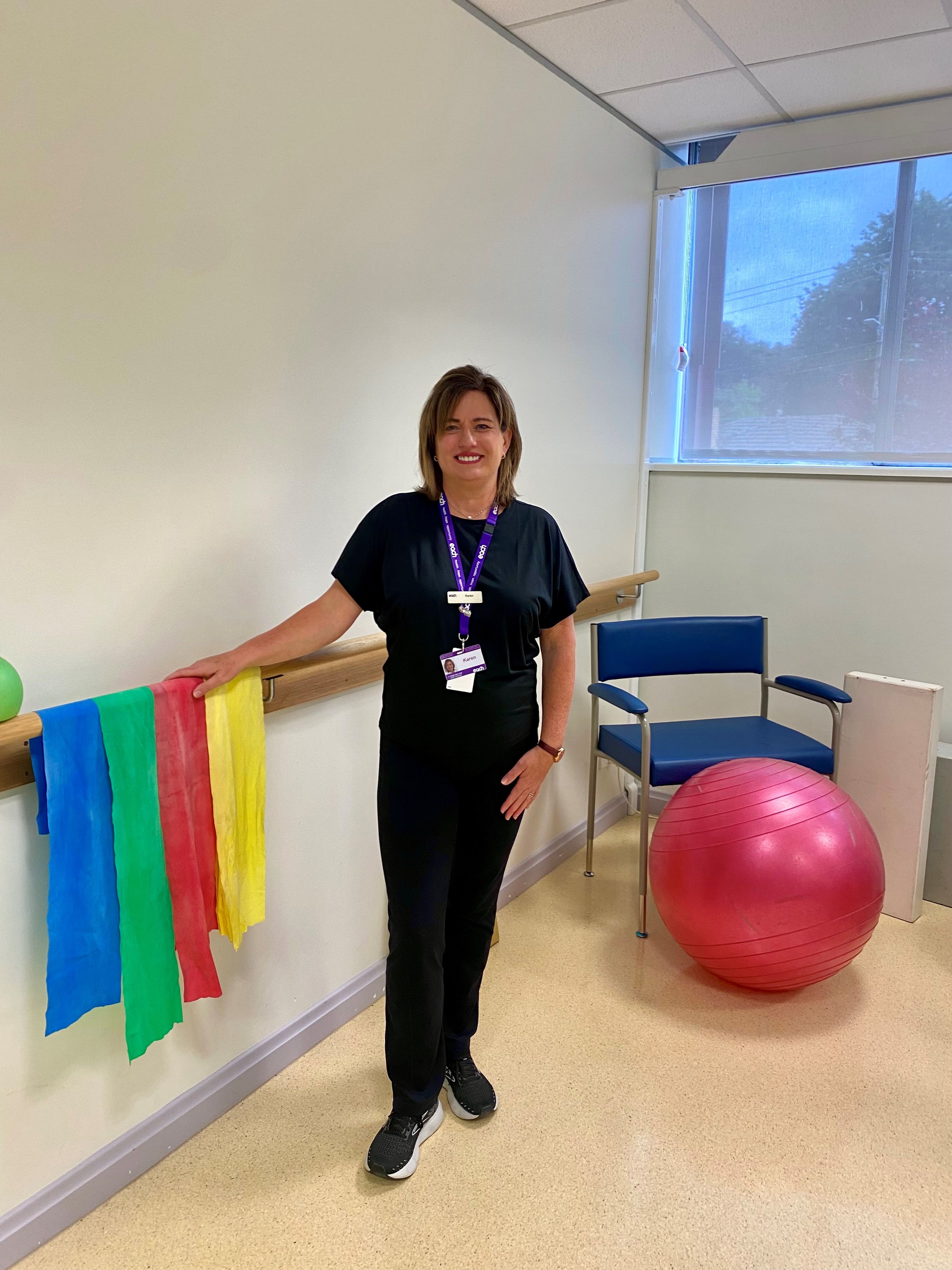 Karen stands in a rehabilitation room, leaning on a handrail with exercise bands handing on it and an exercise ball in the distance.