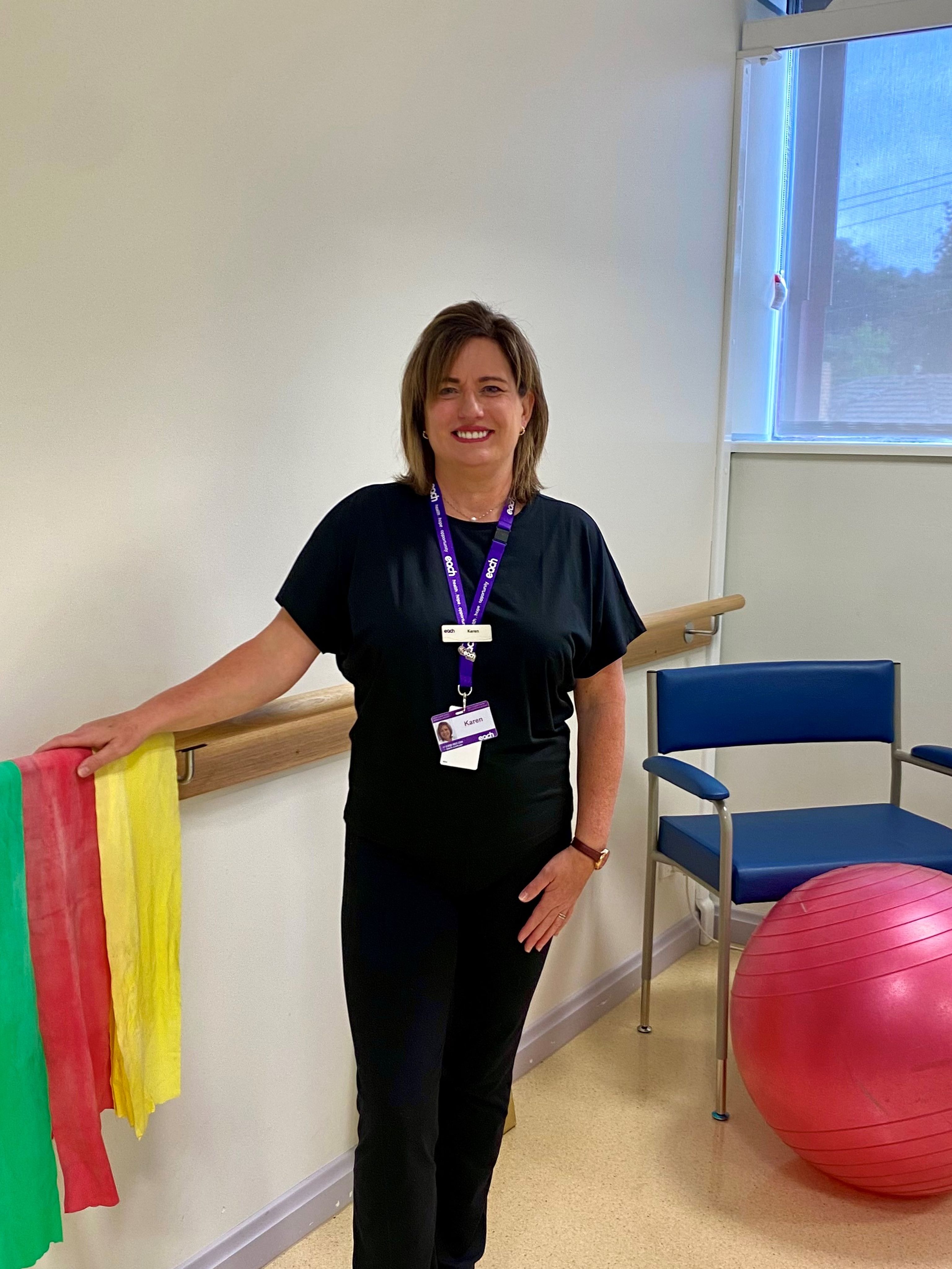 Karen stands in a rehabilitation room, leaning on a handrail with exercise bands handing on it and an exercise ball in the distance.