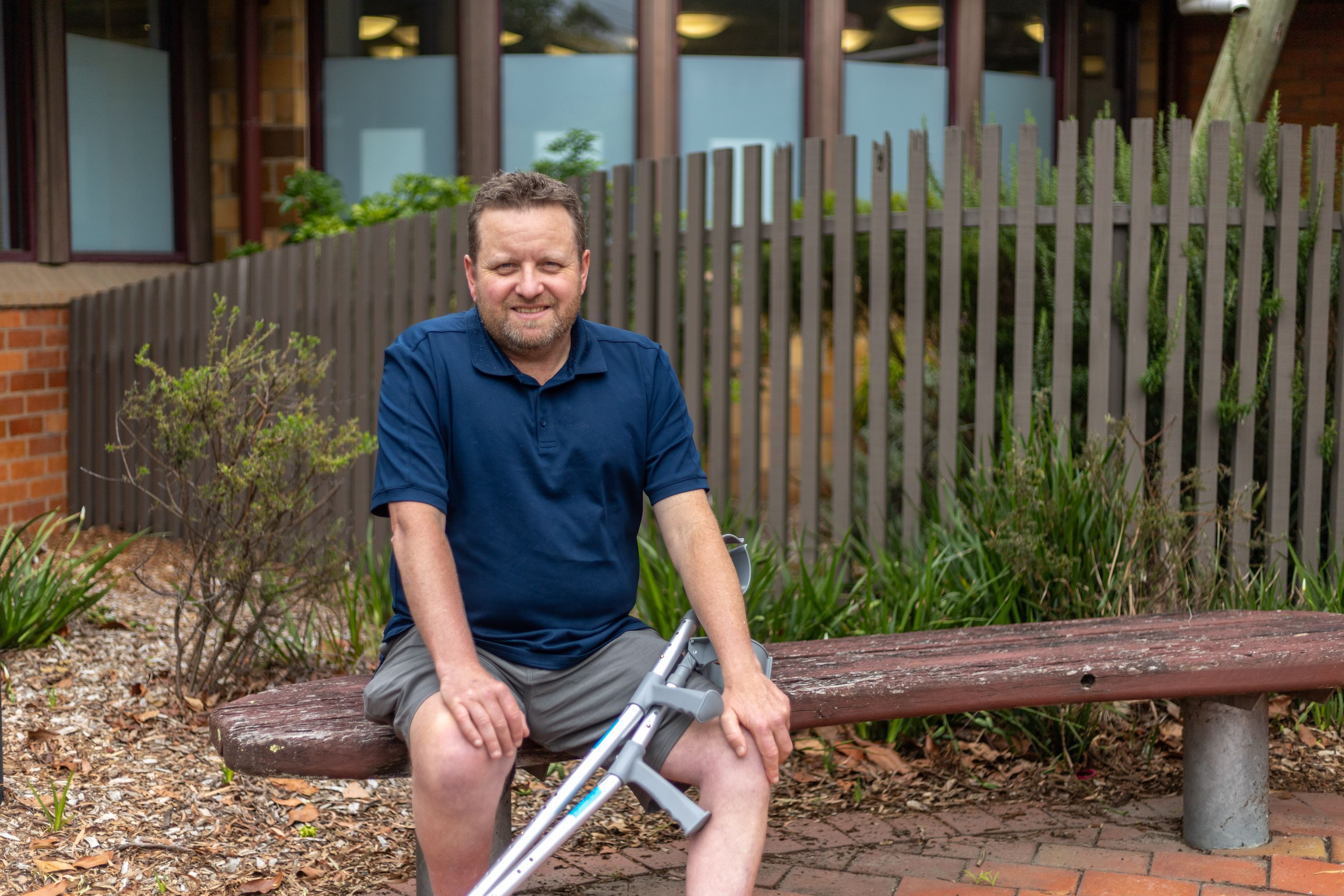 Craig sits on an outdoor bench with a garden behind him. He is smiling. His mobility crutches rest on his knee.