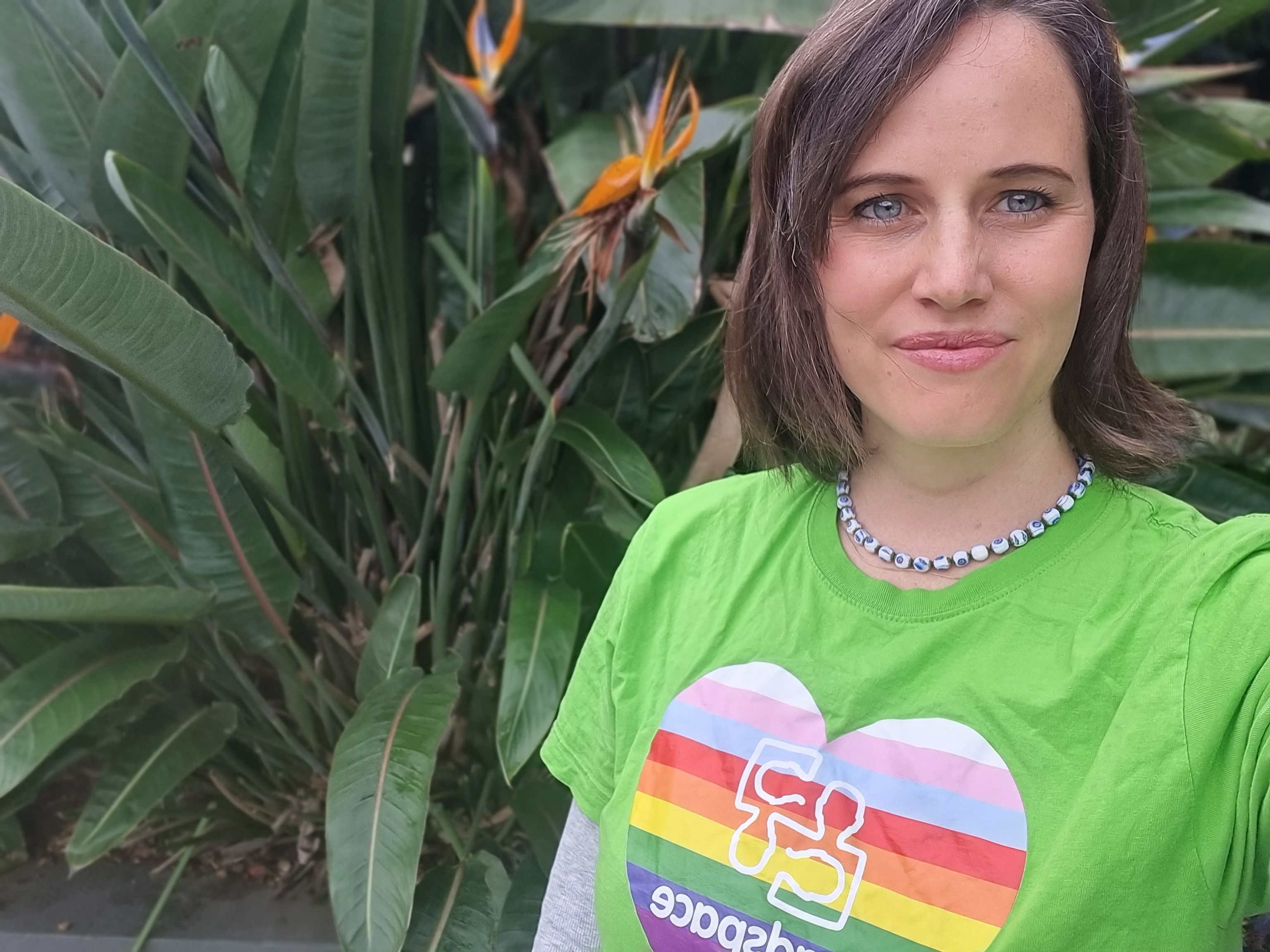Community outreach worker Jenny smiles in front of lush green foliage and bird of paradise flowers.