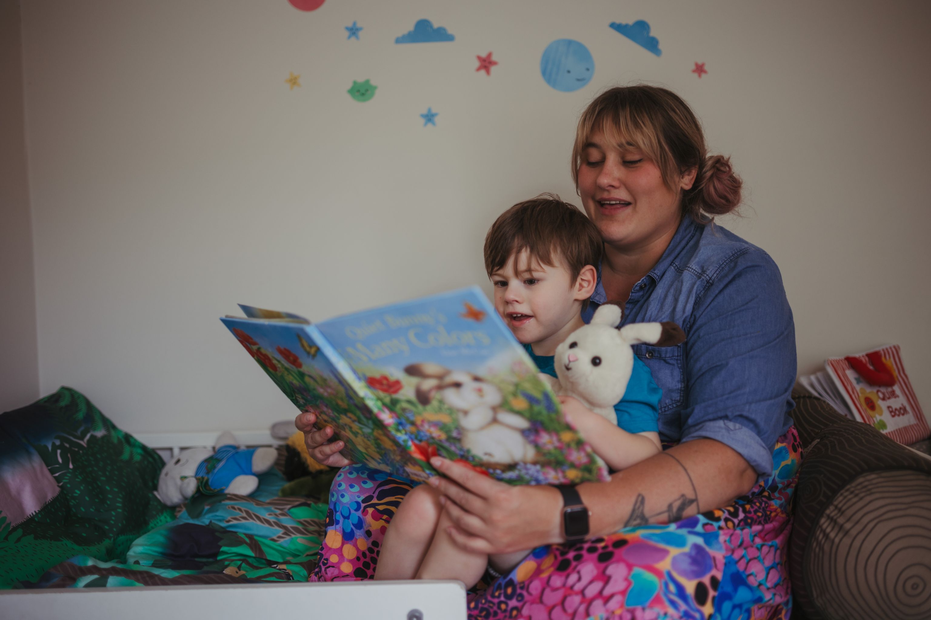 Henry, a young four-year-old boy with dark hair sits on his mother Neah’s lap in a brightly coloured child’s bed. Henry is happily engaged in a story his mum is reading him about a white bunny just like his toy.