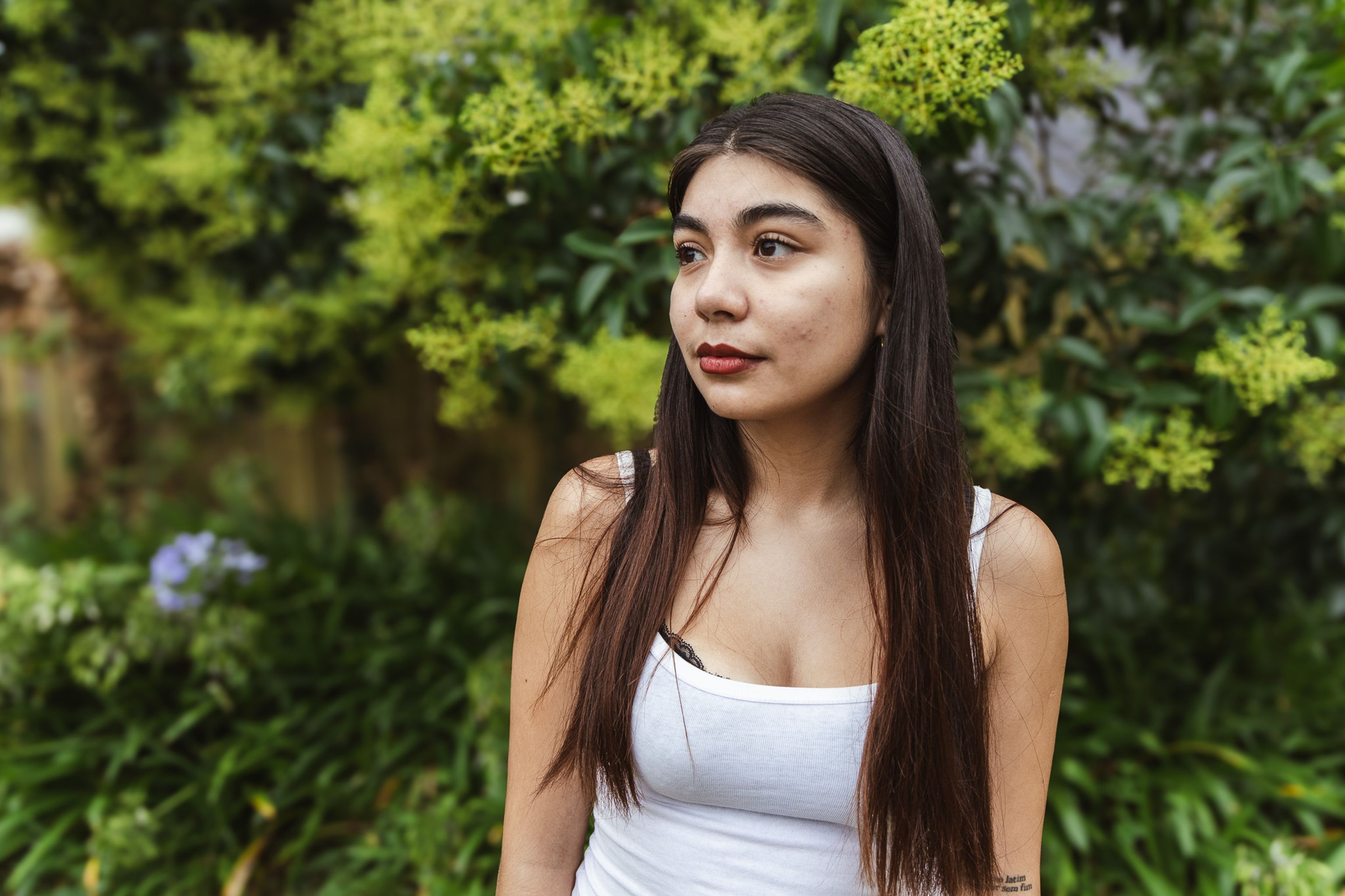 Val, a young woman with long dark hair and a confident expression stands in front of a vibrant blue mural with abstract patterns. She is wearing a white tank top and black jeans, with a tattoo visible on her left arm.